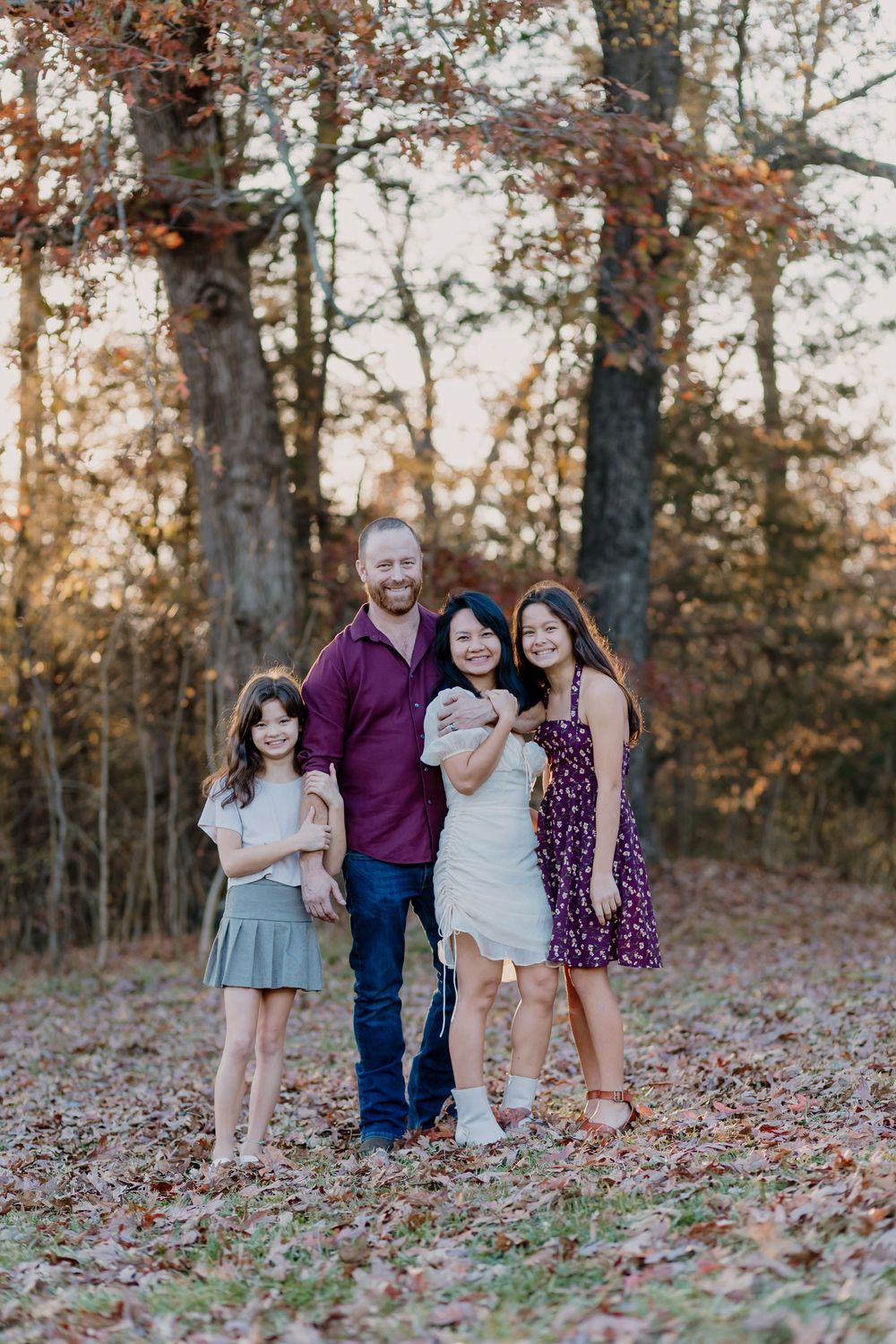 A family of four smiles together in an autumn forest setting, with golden leaves and warm sunset light.