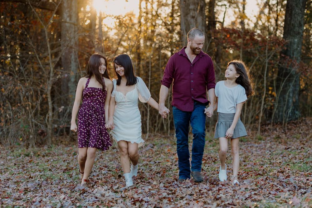 Family of four walking hand-in-hand through autumn woods at golden hour, wearing casual attire.