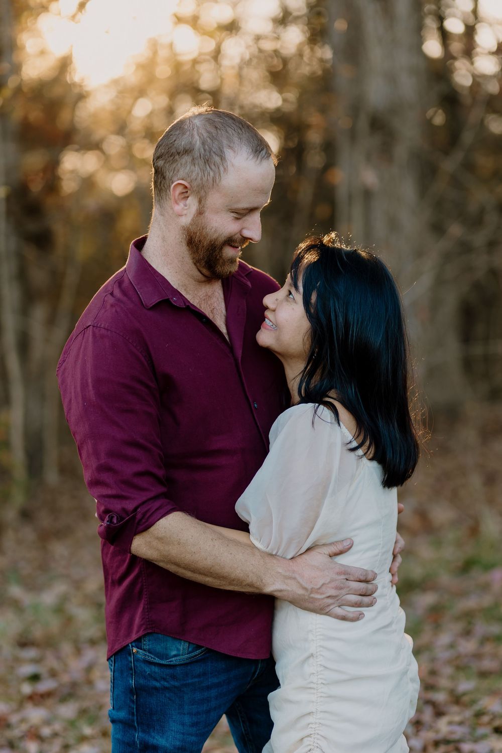 A couple embraces and smiles at each other in a sunlit autumn forest, man in burgundy shirt and woman in white dress.