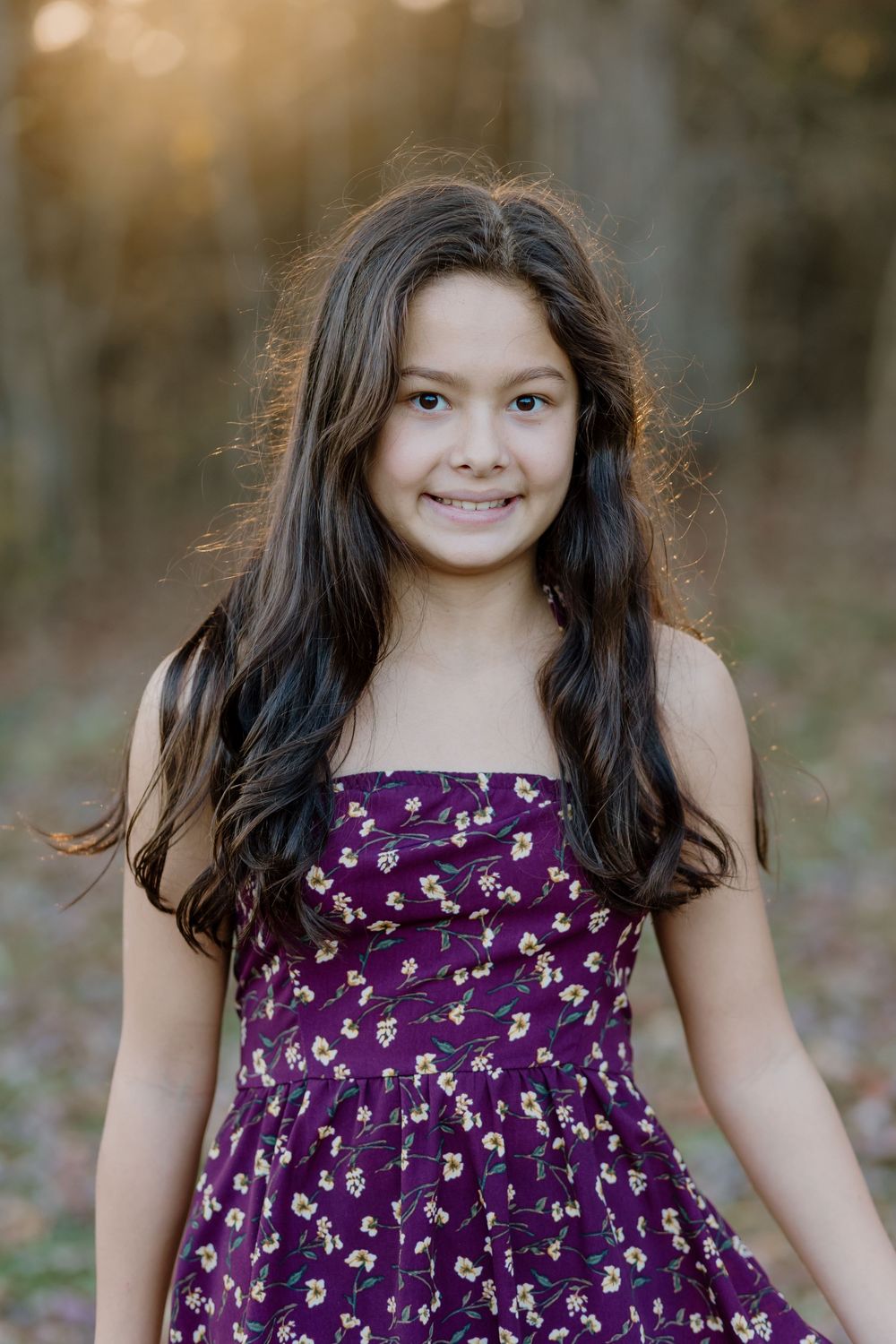 A smiling young girl with long dark hair wearing a purple floral sundress, posed outdoors in a sunlit forest setting.