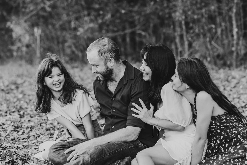 Black and white photo of a happy family of four laughing together outdoors in a wooded setting.