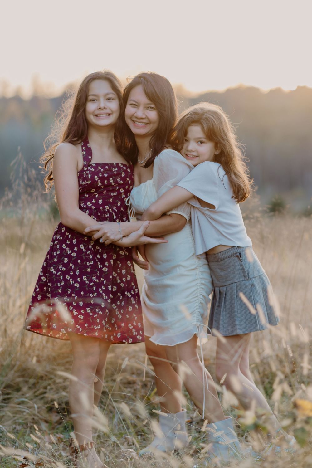 A mother and two daughters embrace in a golden sunlit field, smiling warmly in summer dresses at sunset.