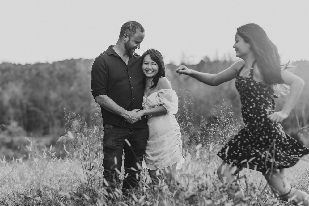 Black and white photo of a couple embracing in a field while a woman in a floral dress dances nearby.