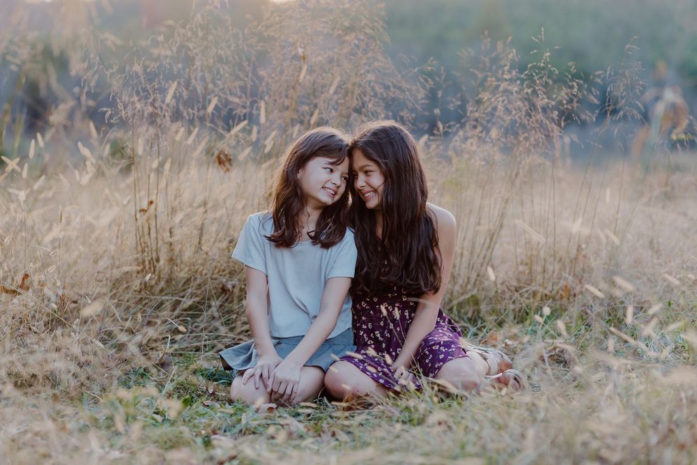 Two young women sitting together in a golden grass field, smiling and leaning on each other in warm sunlight.