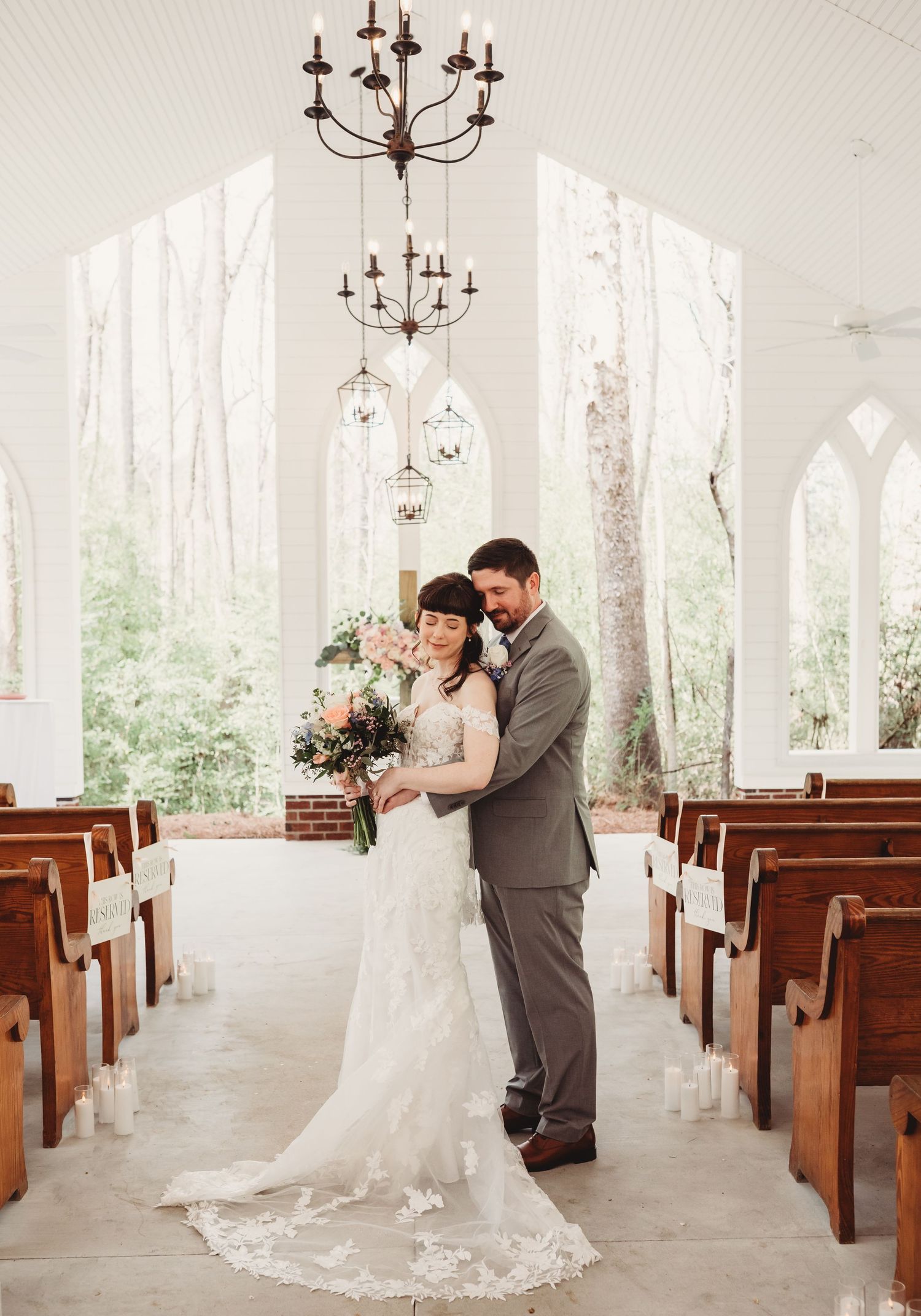 Bride and groom share a tender moment in a white chapel aisle, surrounded by wooden pews and a chandelier.