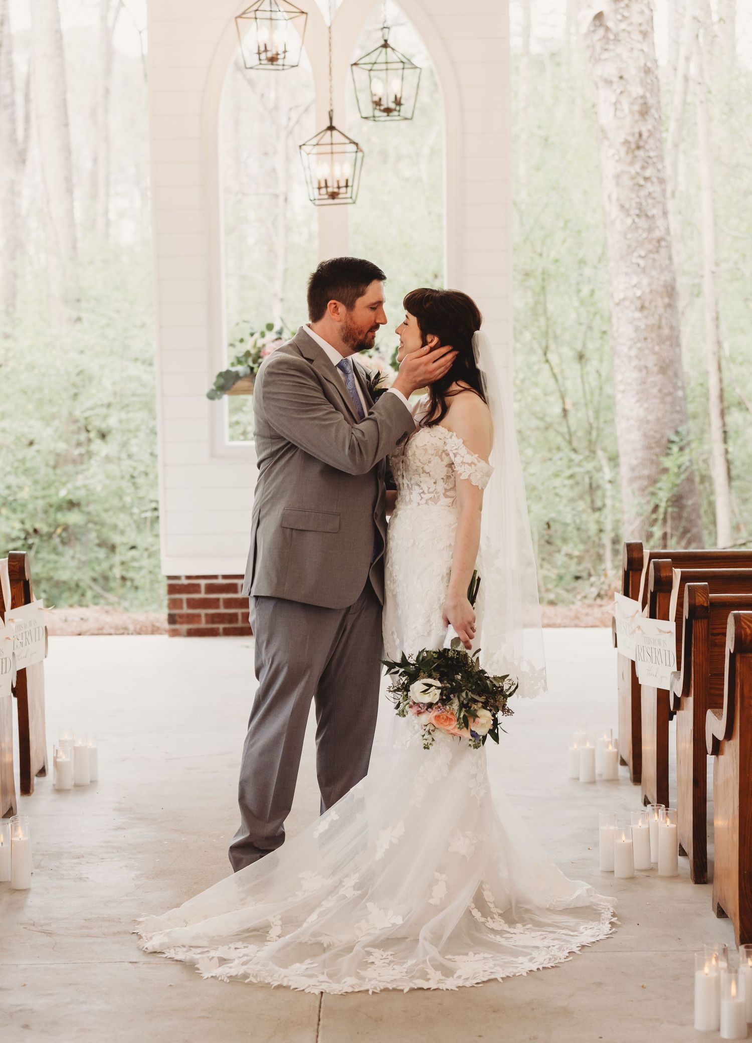 Bride and groom share a romantic moment at the altar of a chapel with wooden pews, candles, and hanging lanterns.