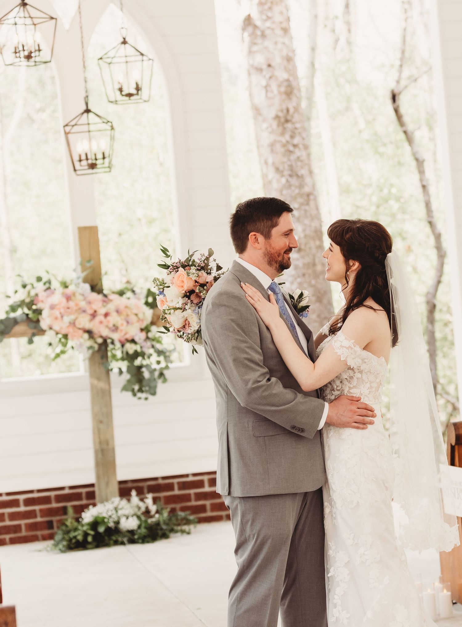 A bride and groom share a romantic moment in an elegant white chapel with lanterns and pink floral arrangements.