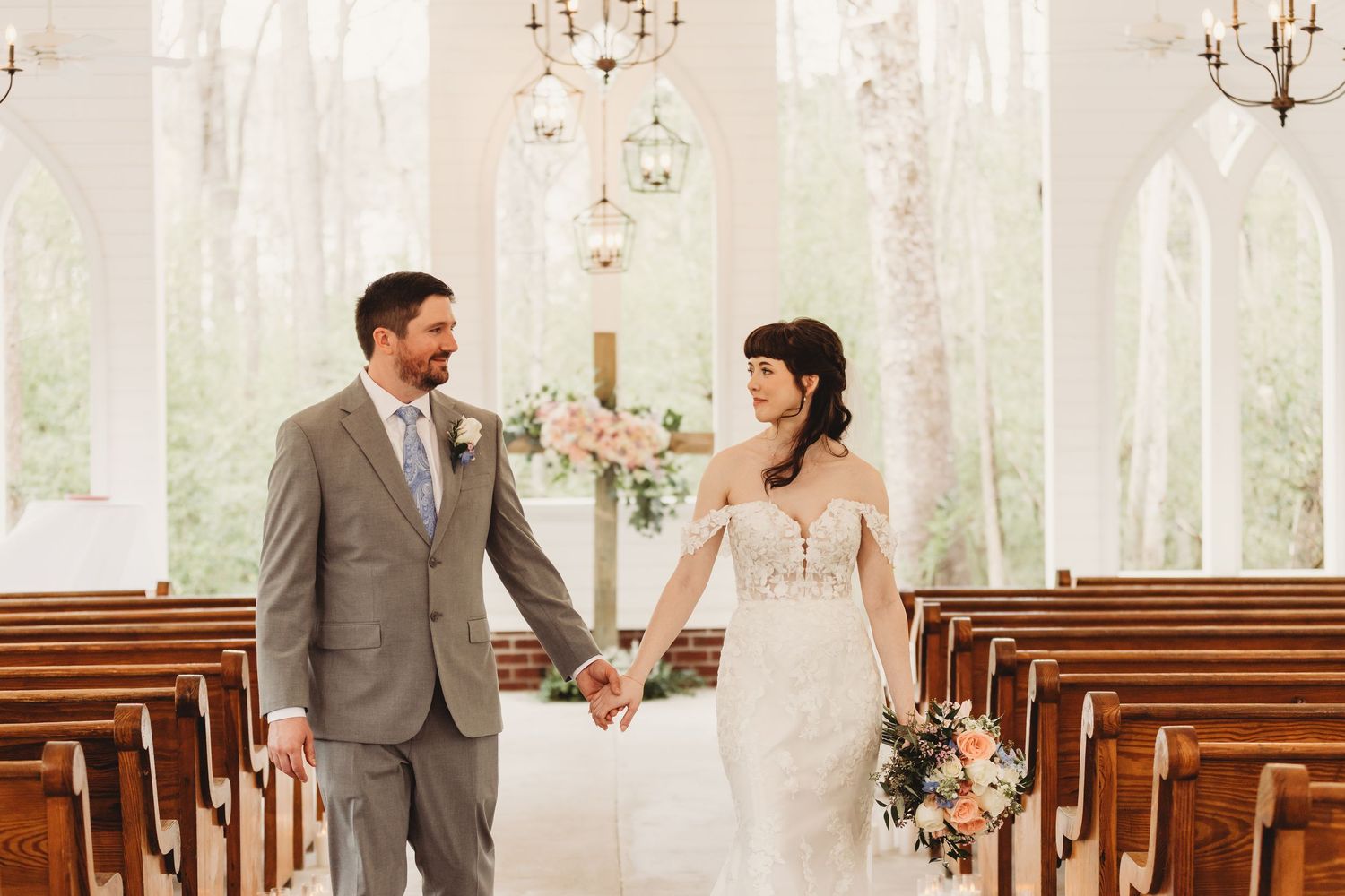 Bride and groom hold hands smiling at each other in a bright white chapel with wooden pews and chandelier lighting.