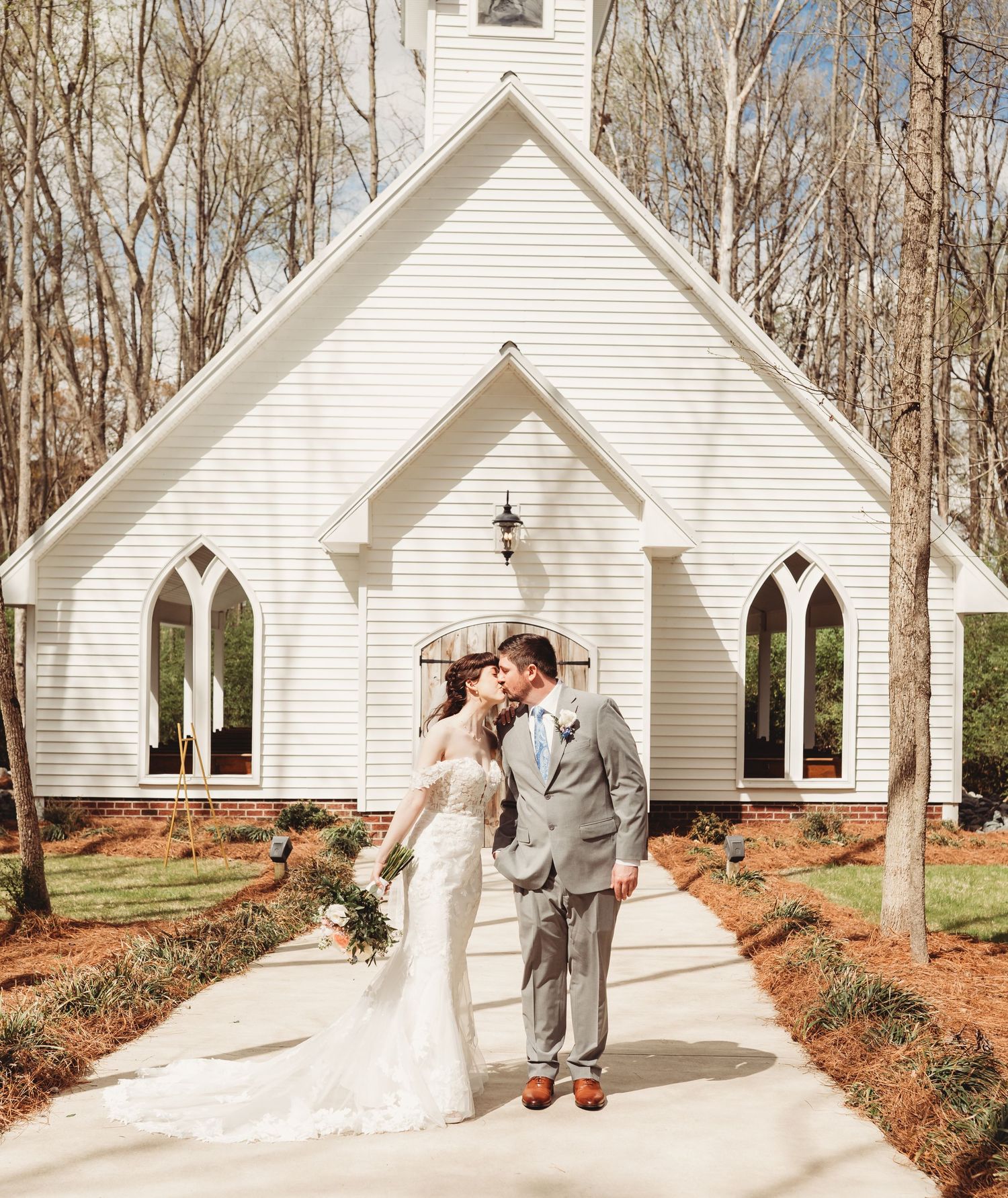 Bride and groom kissing in front of a white chapel surrounded by trees on their wedding day.