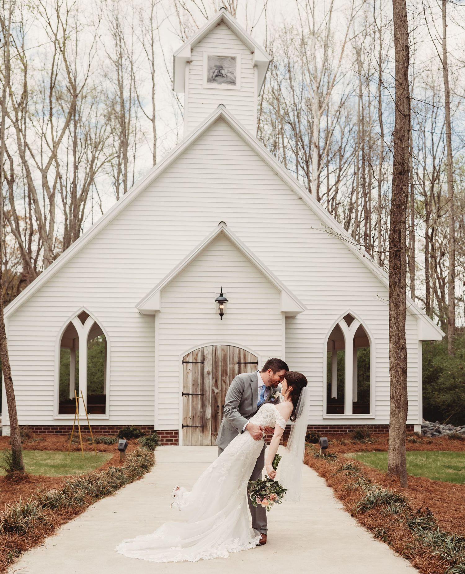 Bride and groom share a romantic dip kiss in front of a charming white chapel surrounded by trees.