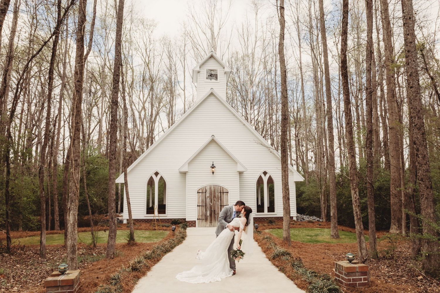 Bride and groom embrace on a chapel path surrounded by tall trees at a romantic outdoor wedding venue.