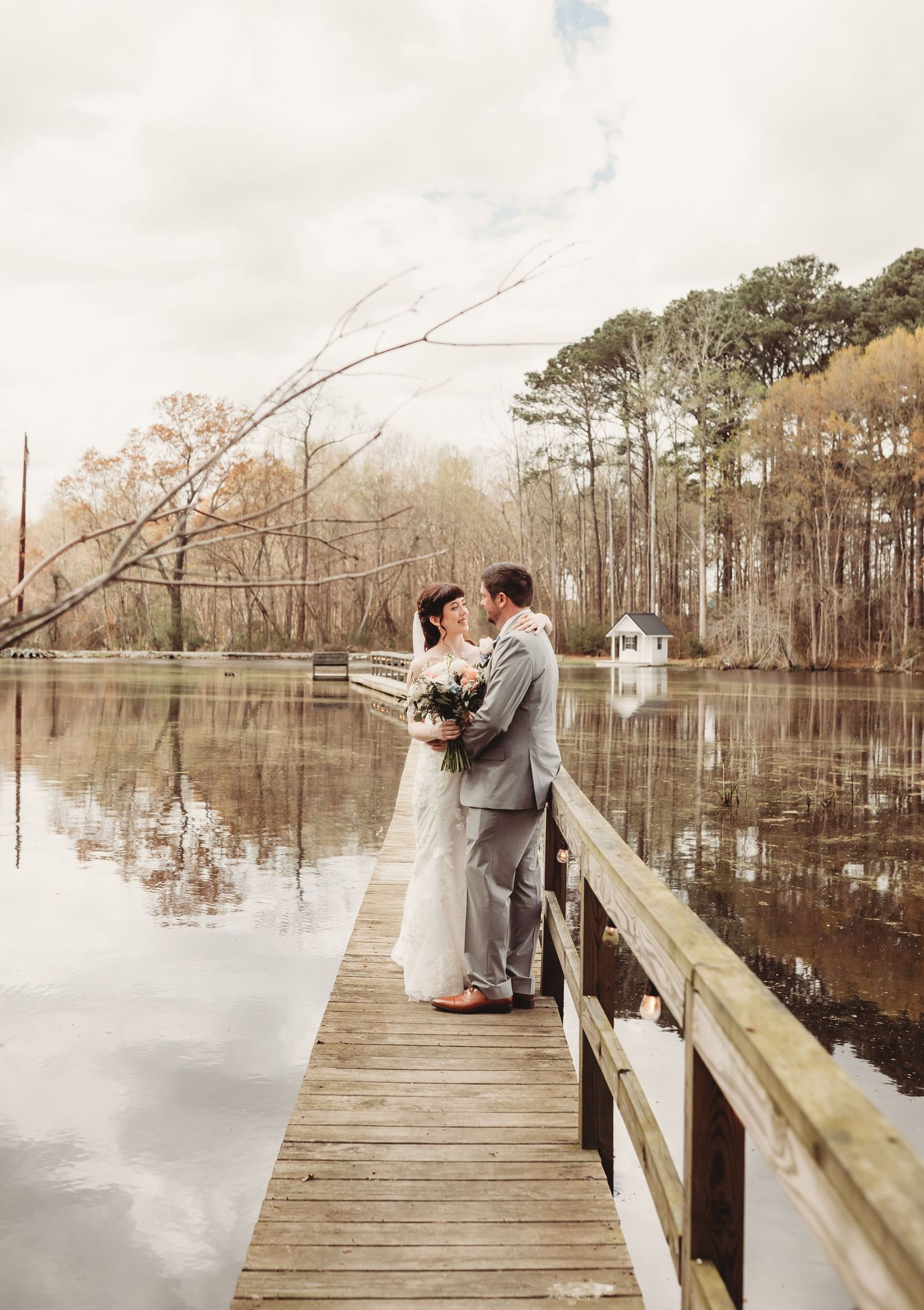 Bride and groom share a romantic moment on a wooden dock overlooking a serene lake surrounded by autumn trees.