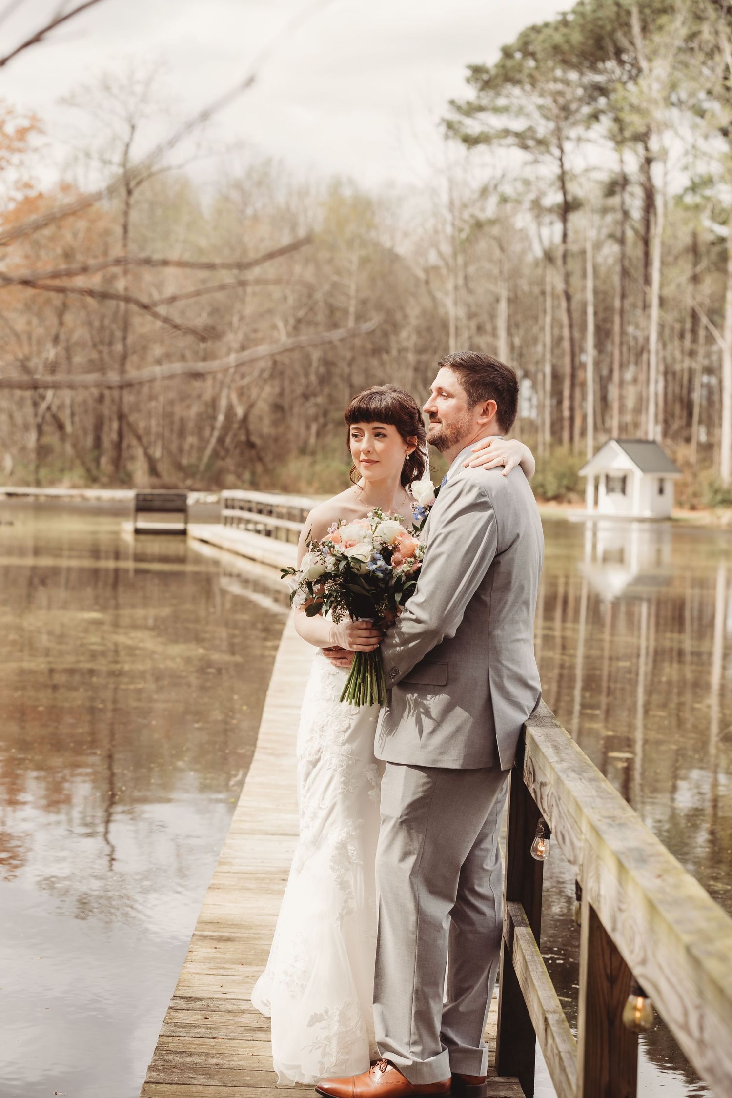 Bride and groom embrace on a wooden dock by a serene lake, surrounded by autumn trees, bride holding a bouquet.
