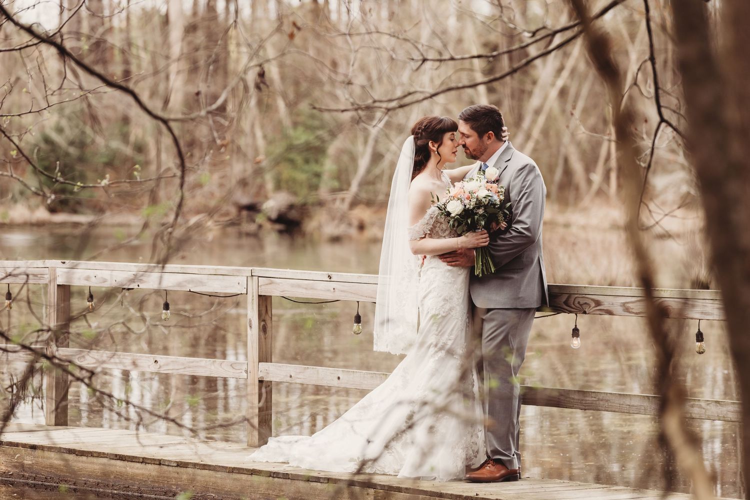 Bride and groom kissing on a wooden dock surrounded by bare trees and a serene pond on their wedding day.