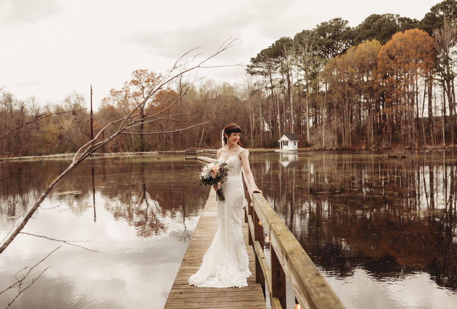 Bride in white lace gown holding bouquet on wooden dock over calm lake surrounded by autumn trees.