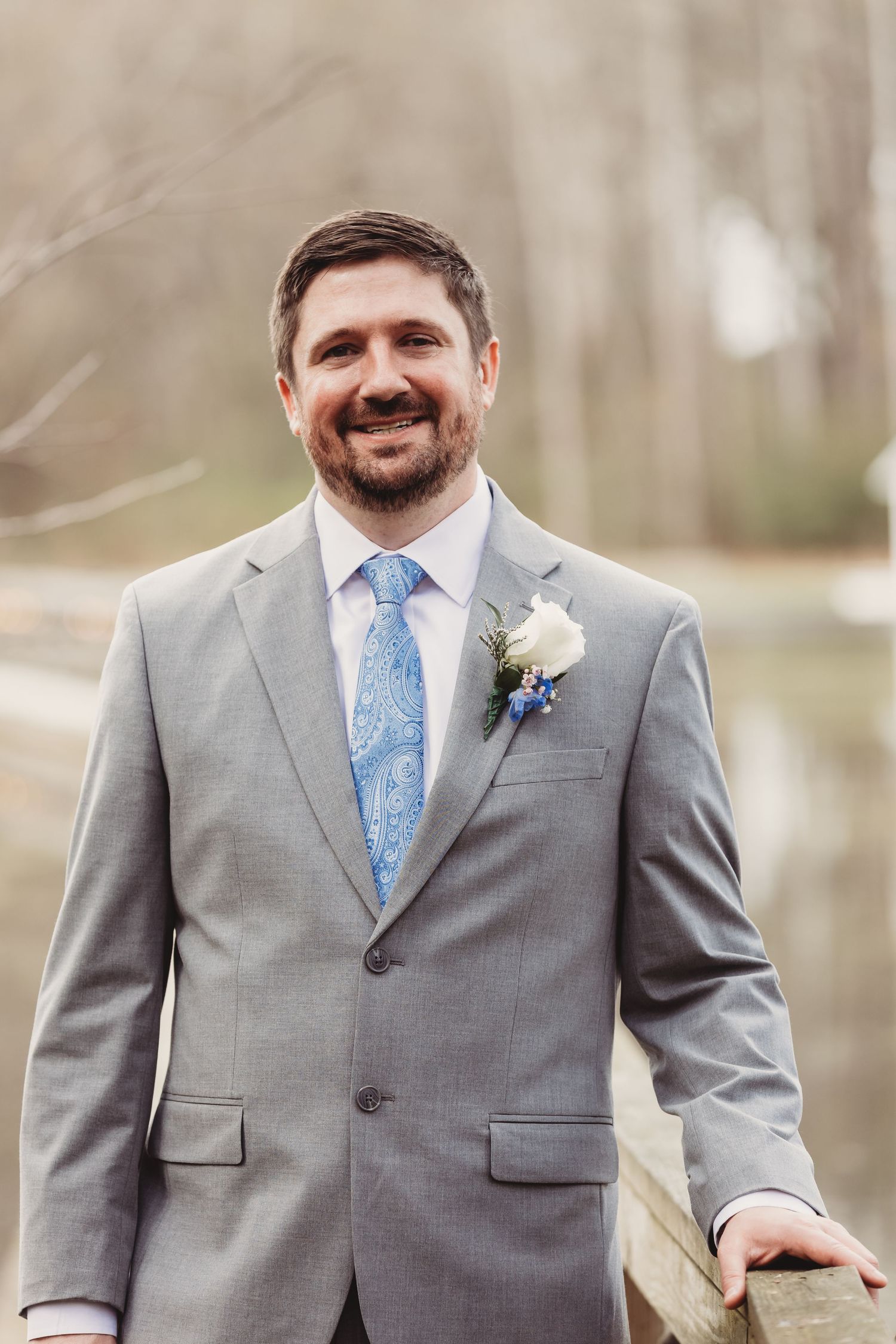 Smiling groom in gray suit with blue paisley tie and white boutonniere posing outdoors at wedding.