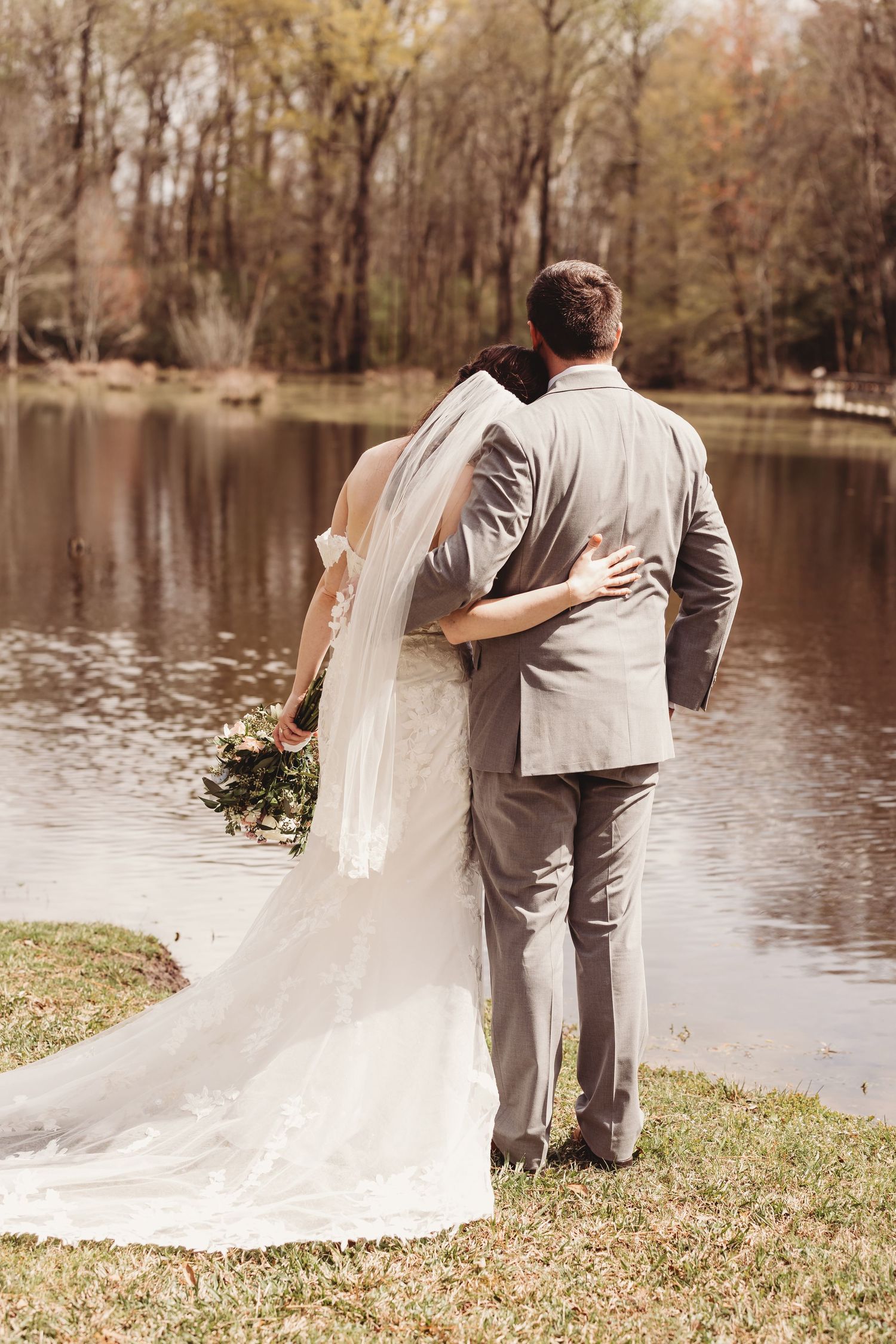 Bride and groom embracing by a serene lake, bride holding floral bouquet with long veil trailing on green grass.