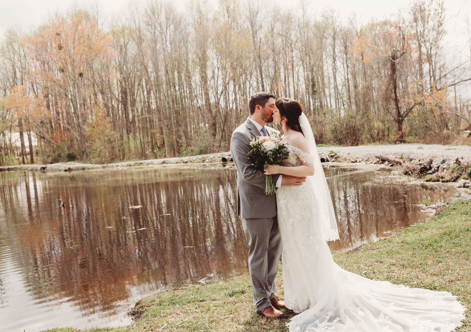 Bride and groom share a romantic kiss by a serene pond surrounded by autumn trees on their wedding day.