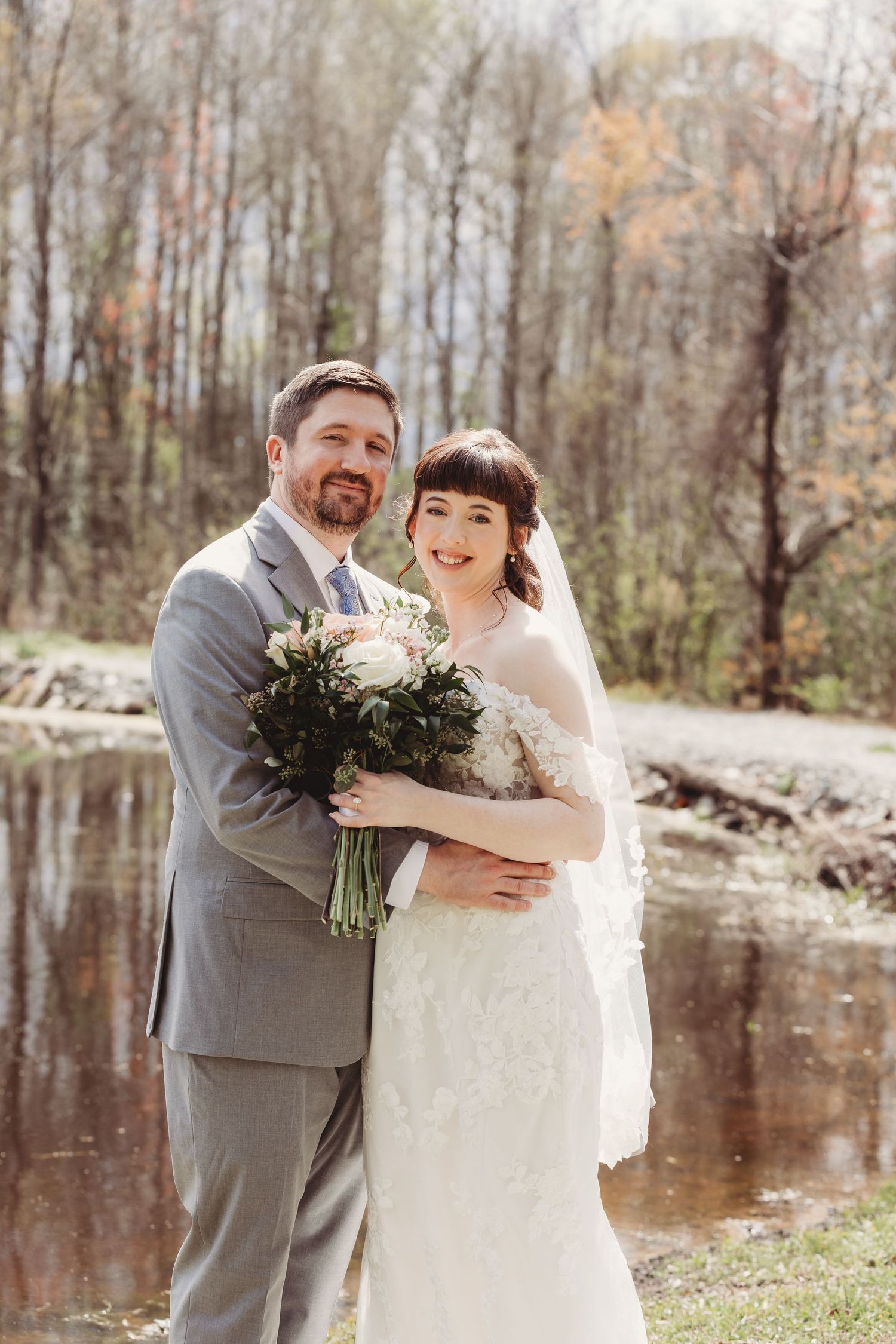 A bride and groom pose together outdoors near a pond, she holds a bouquet in a lace gown with veil.