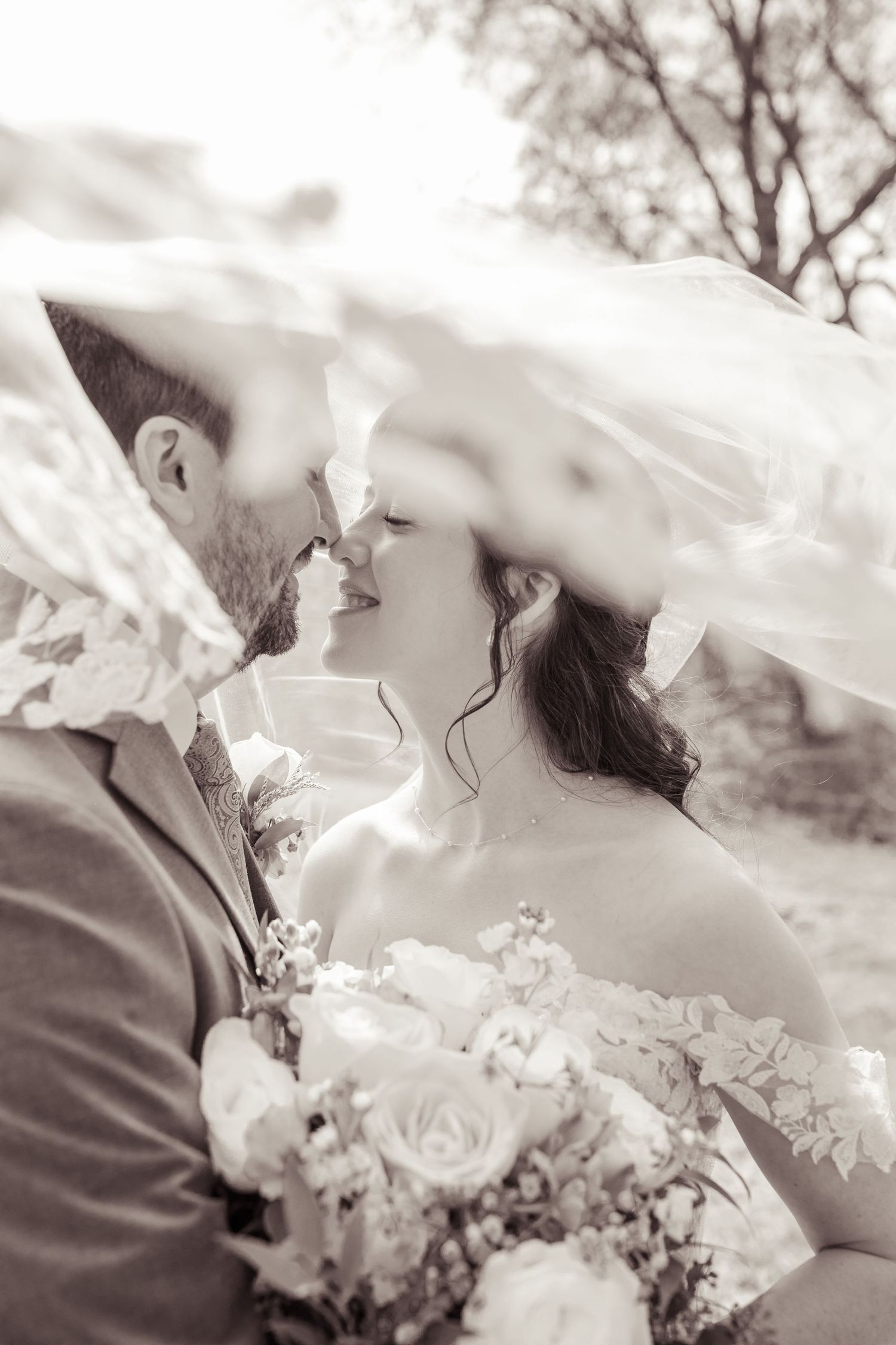 Black and white photo of bride and groom almost kissing under flowing veil, bride holding roses bouquet outdoors.