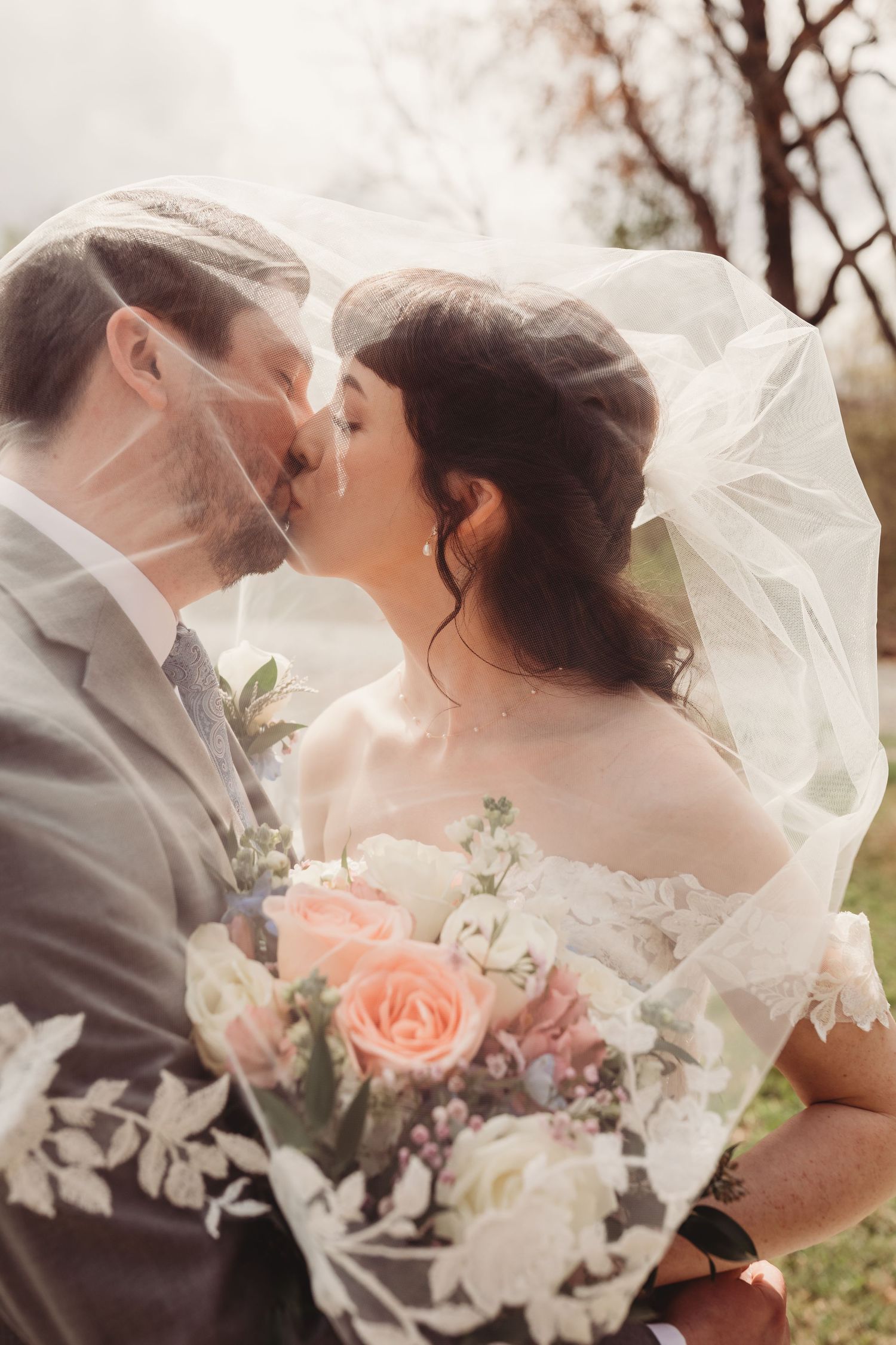 Bride and groom kissing under a lace wedding veil, bride holding a bouquet of pink and white roses outdoors.