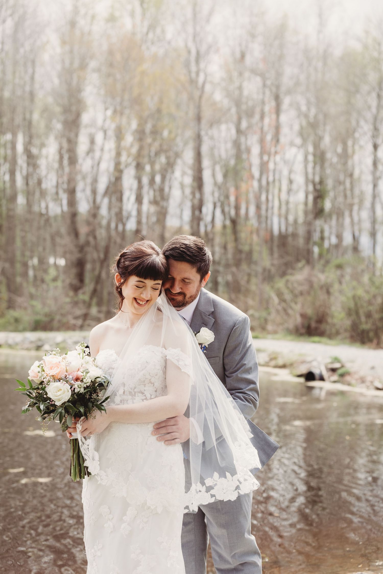 Bride and groom embrace near a serene forest creek, bride holding a blush bouquet in a lace gown with veil.