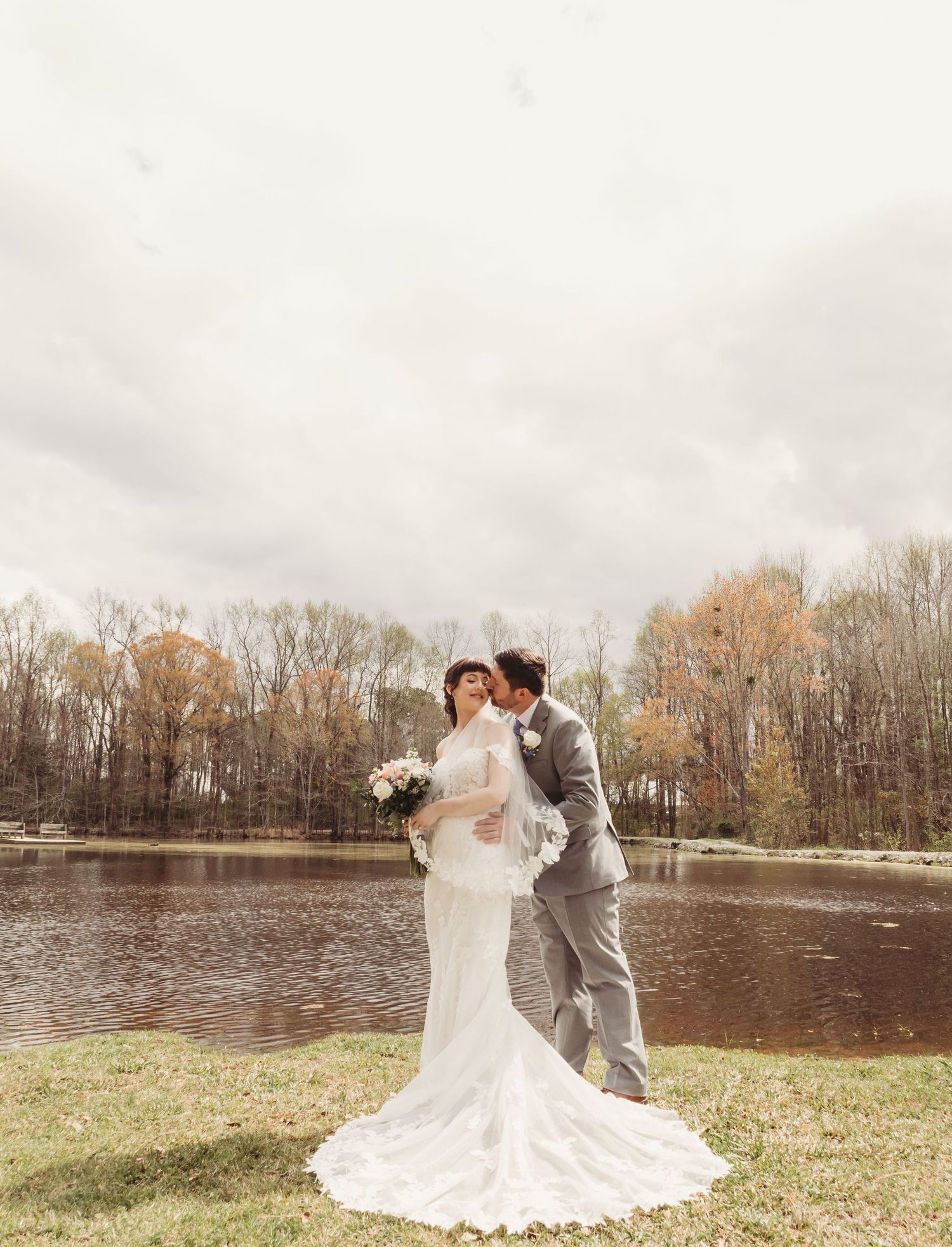 Bride and groom share a romantic kiss by a serene lake surrounded by autumn trees on their wedding day.