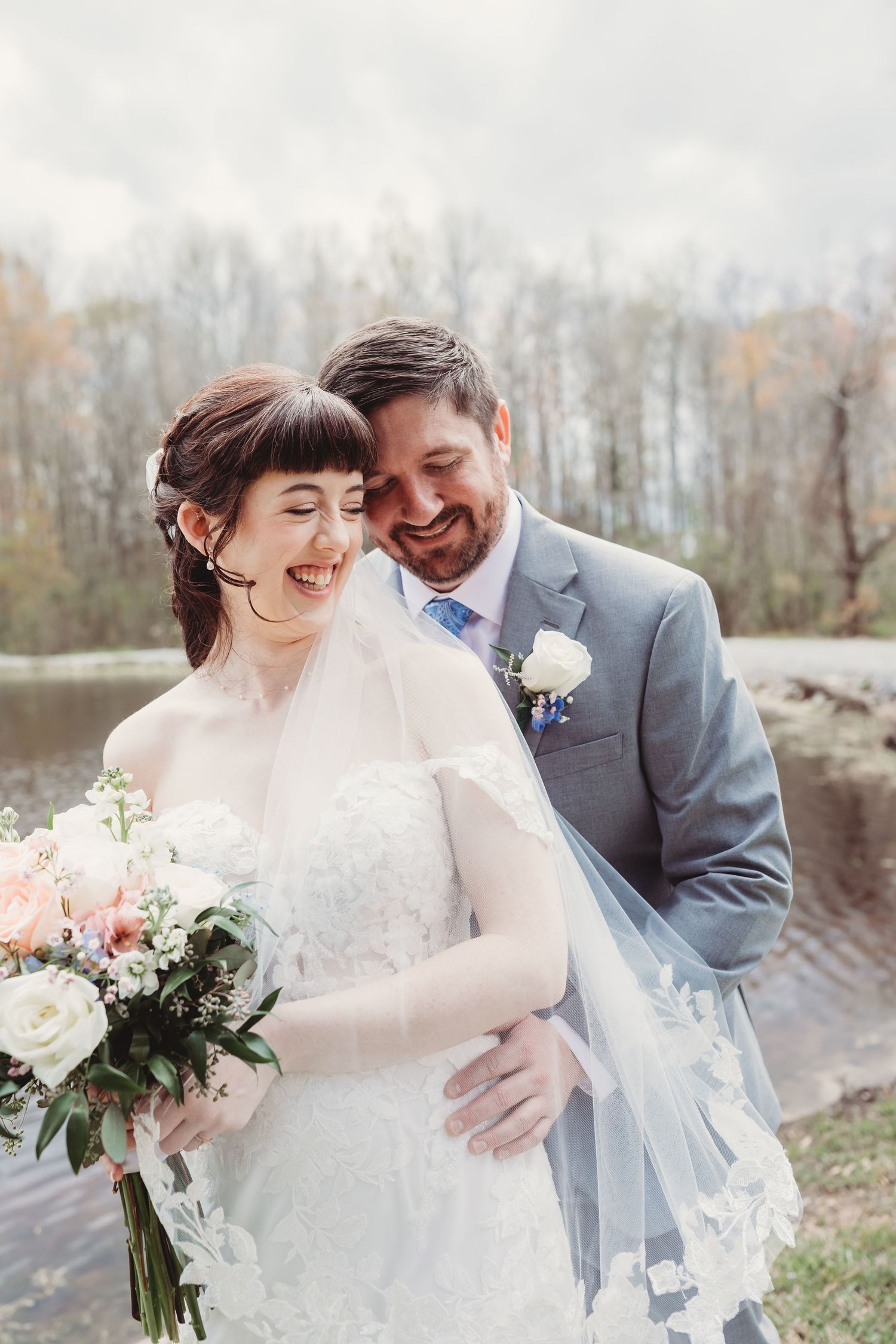 Smiling bride and groom embracing outdoors near a lake, bride holding a white and pink bouquet, groom in gray suit.