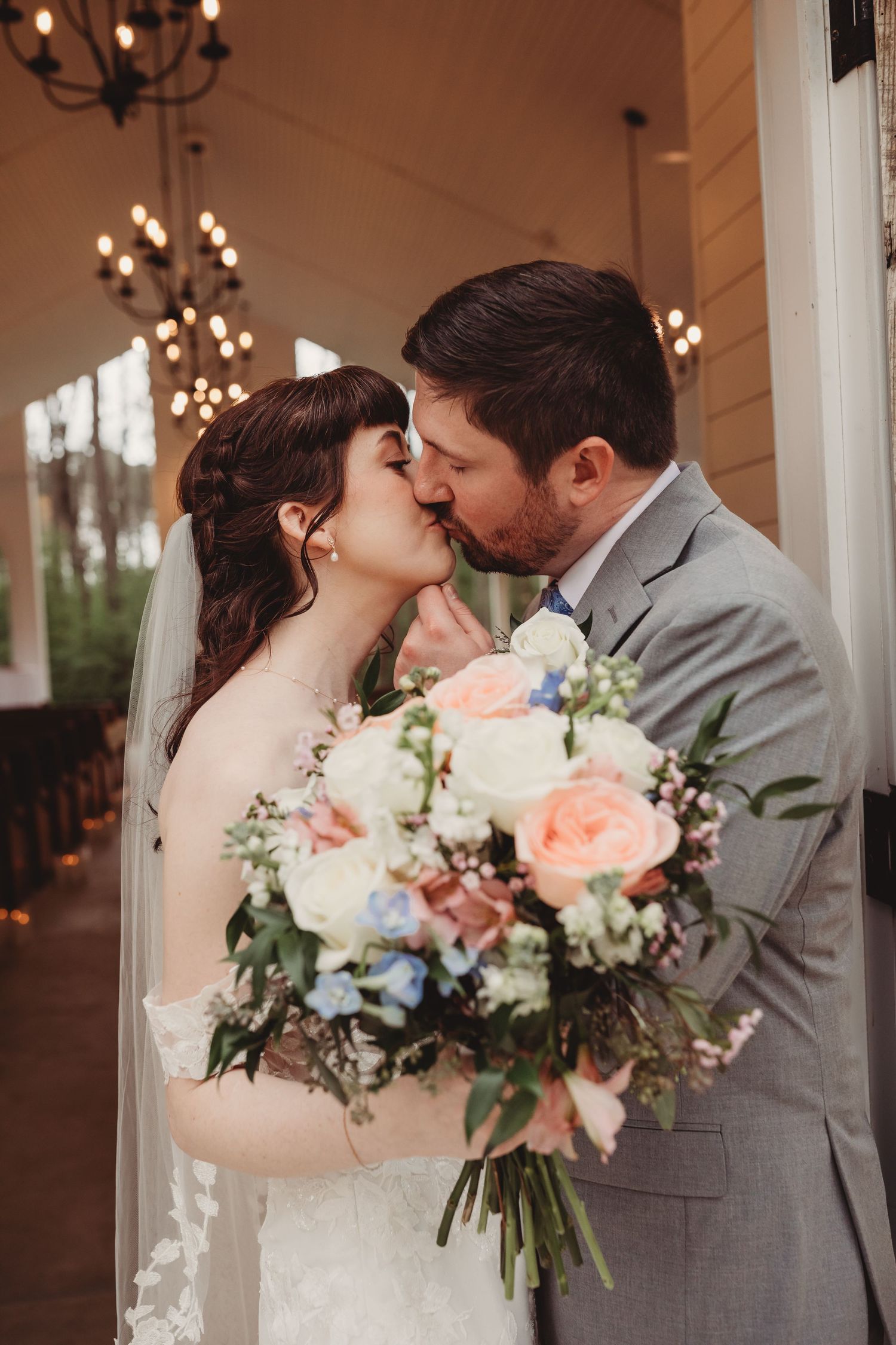 Bride and groom share a kiss while bride holds a lush bouquet of peach, white, and blue flowers at elegant venue.