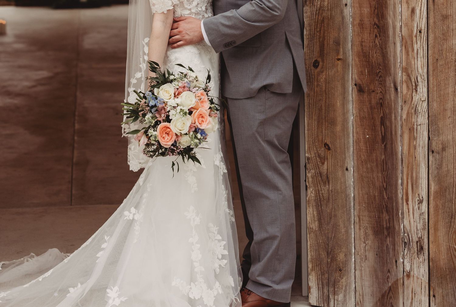 A bride in a lace gown holds a peach floral bouquet beside a groom in a gray suit near a rustic wooden wall.