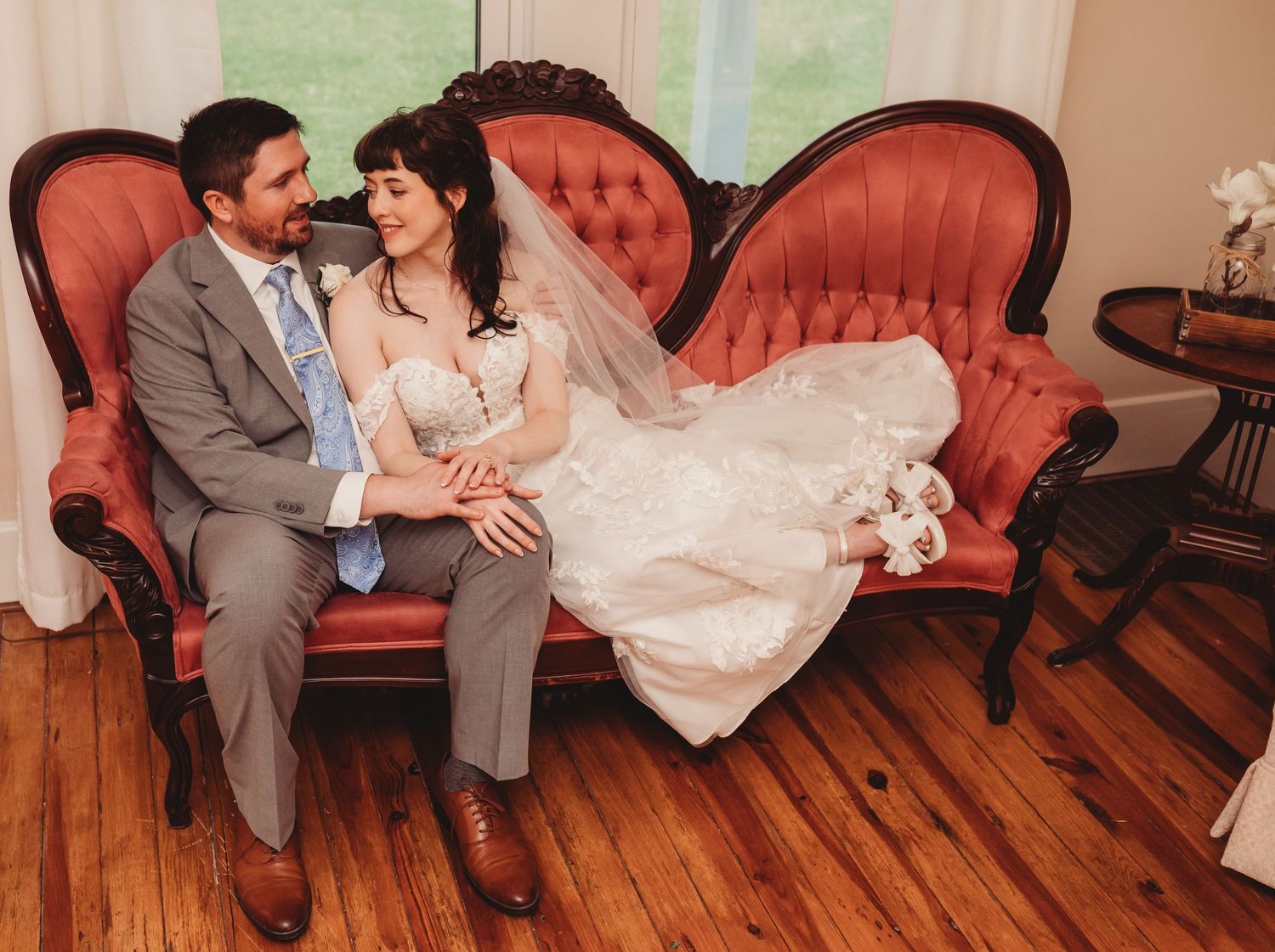 Bride and groom sitting together on a vintage red velvet sofa, sharing a tender moment in their wedding attire.