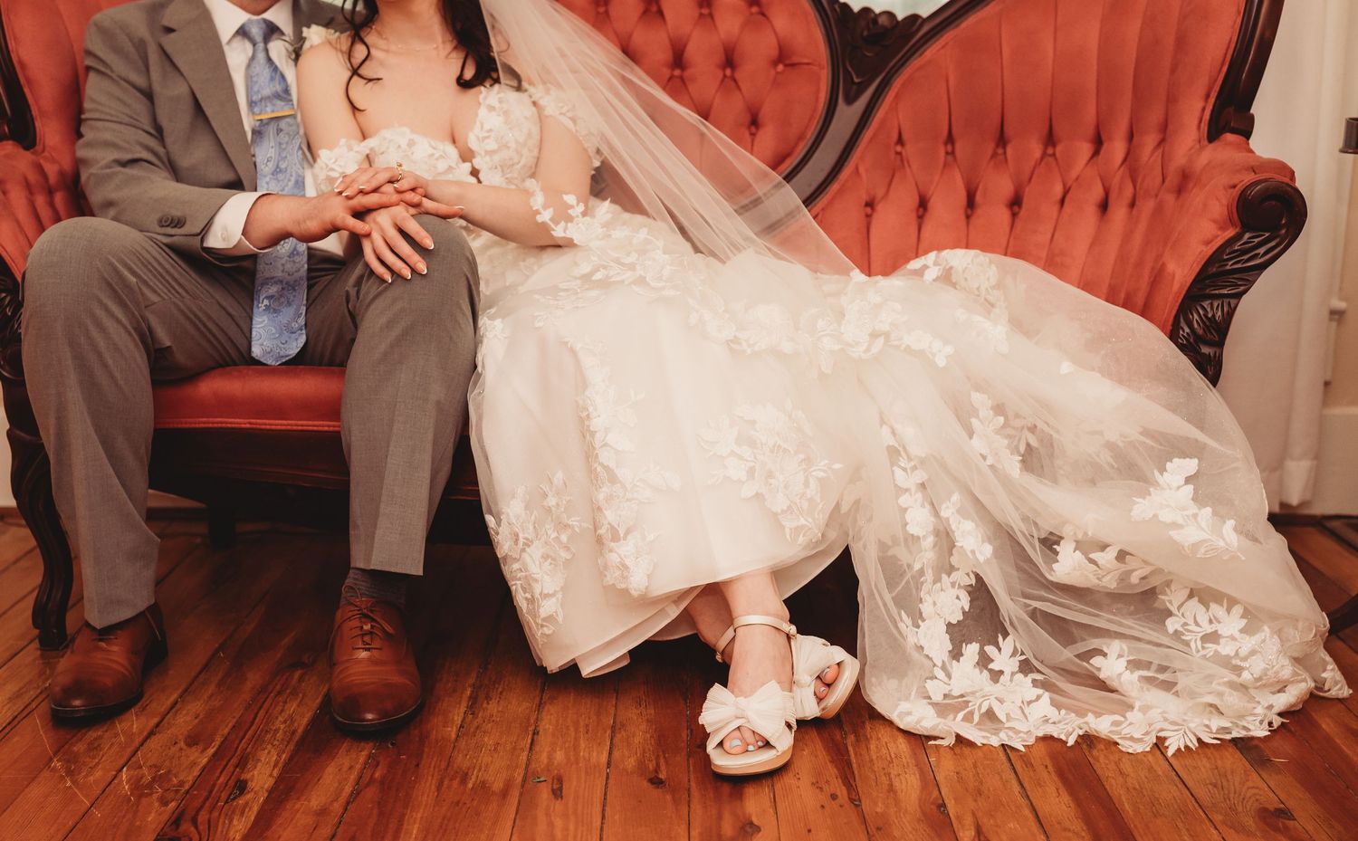 Bride and groom sitting on a vintage red velvet sofa, bride wearing lace wedding dress with veil and white heels.