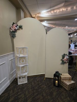 Elegant wedding backdrop with two ivory arch panels adorned with blush floral arrangements, LOVE letter boxes, lantern, and wooden crate.