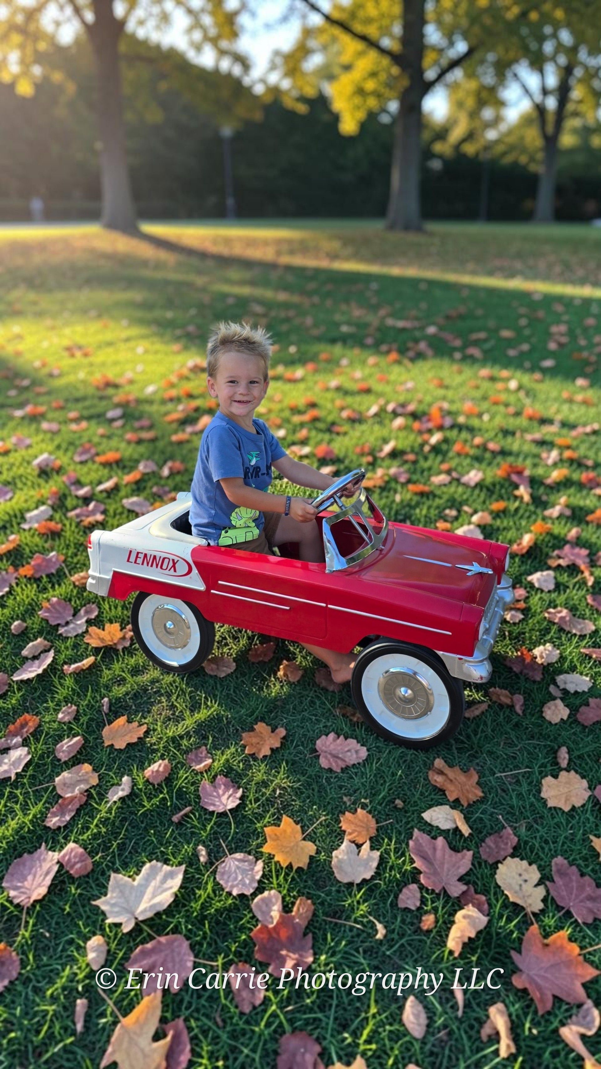 Smiling toddler boy sitting in a red vintage Lennox pedal car on a grass lawn covered with colorful autumn leaves.