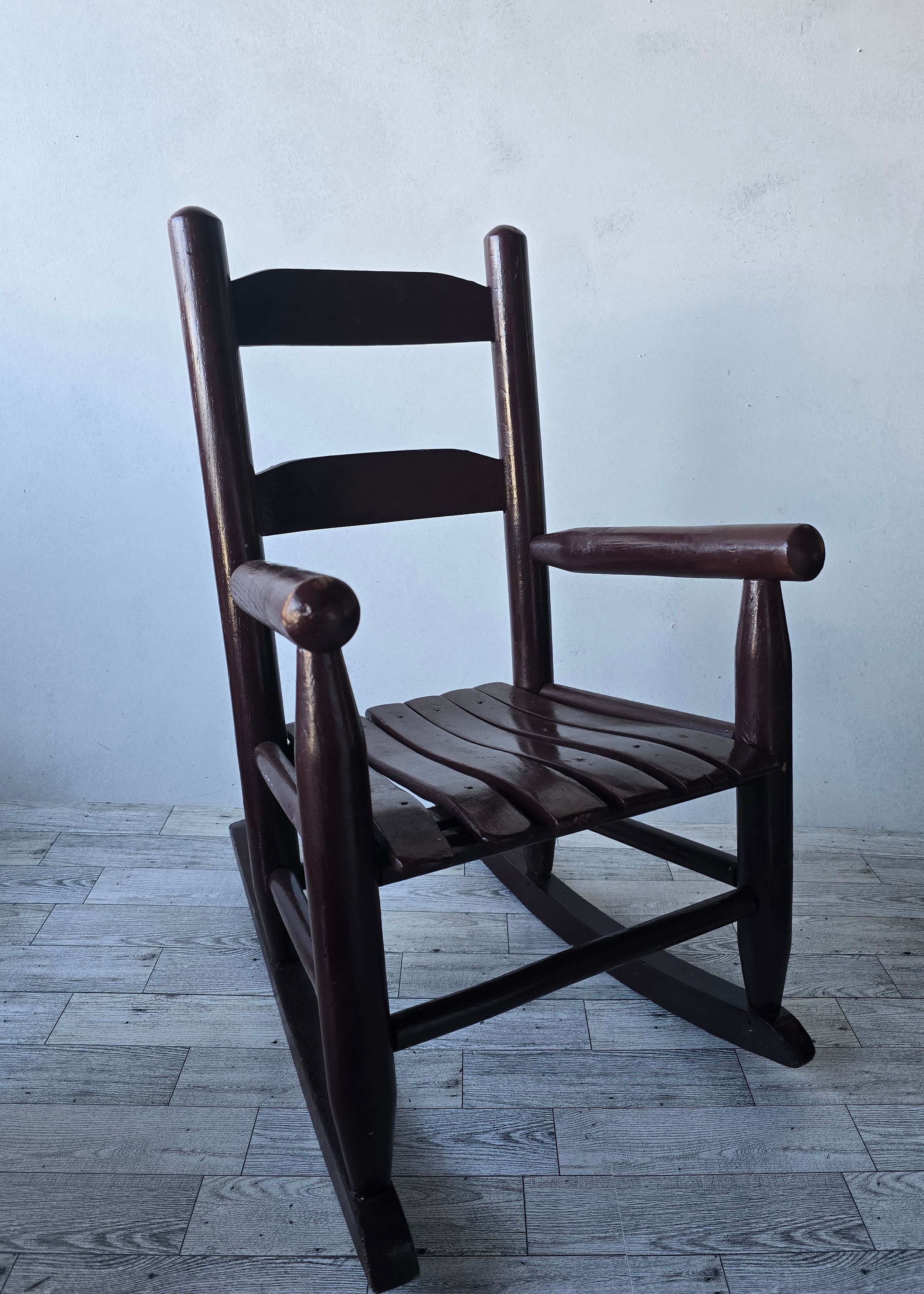 Dark wood child's rocking chair with slatted seat and ladder back design, displayed against a light gray wall.