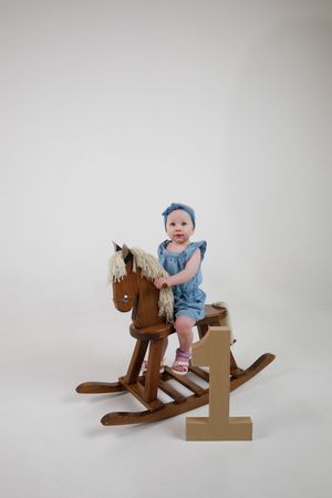 Baby girl in blue romper sitting on wooden rocking horse next to number 1 block on white background.