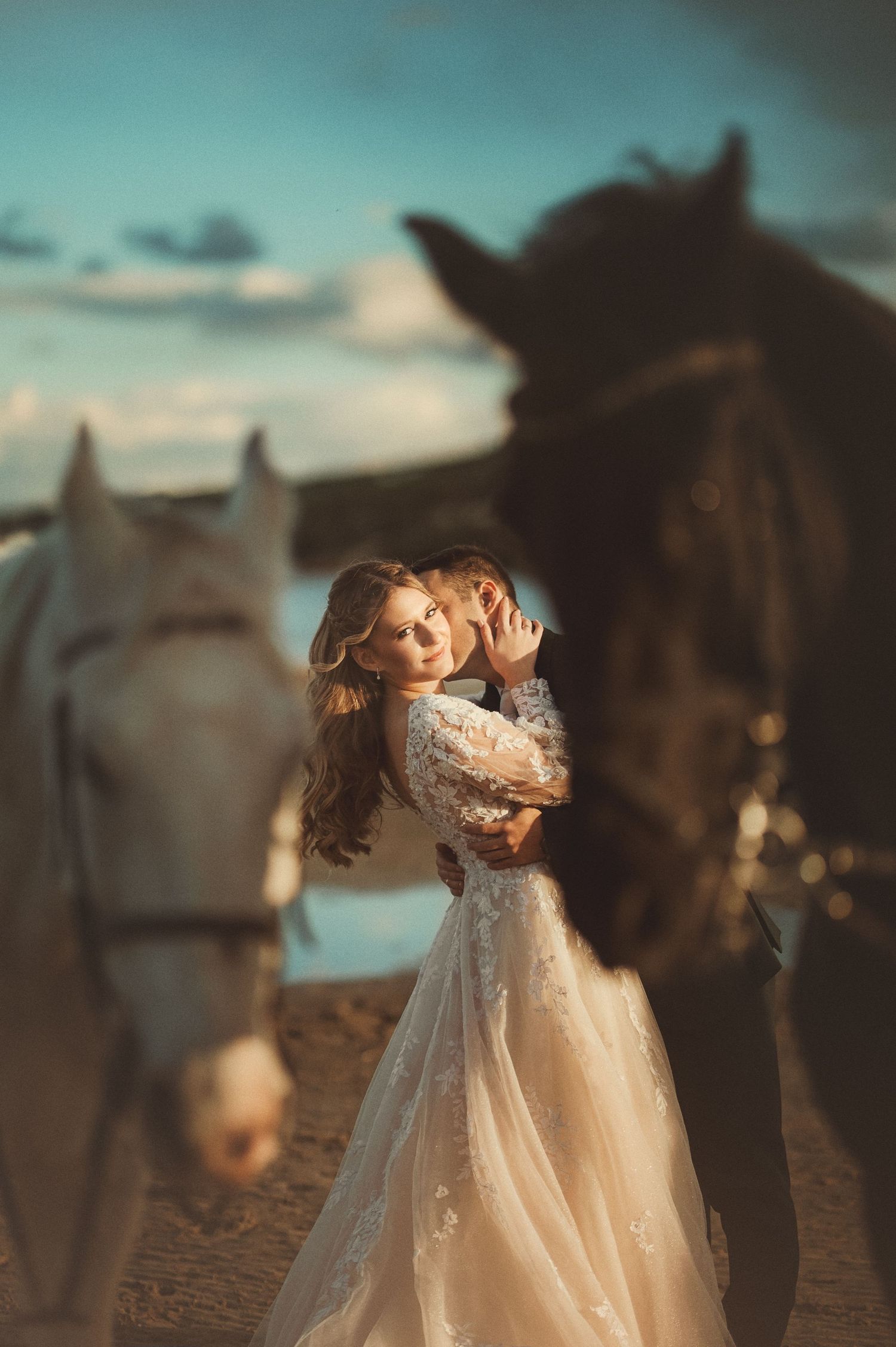 A bride in a lace gown embraces her groom between a white and dark horse outdoors.
