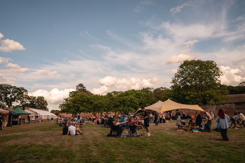 Outdoor festival gathering with tents, crowds on green lawn, trees, and blue sky with clouds at golden hour.