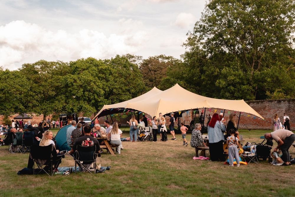 Outdoor community gathering under a large stretch tent on green lawn, surrounded by trees at dusk.