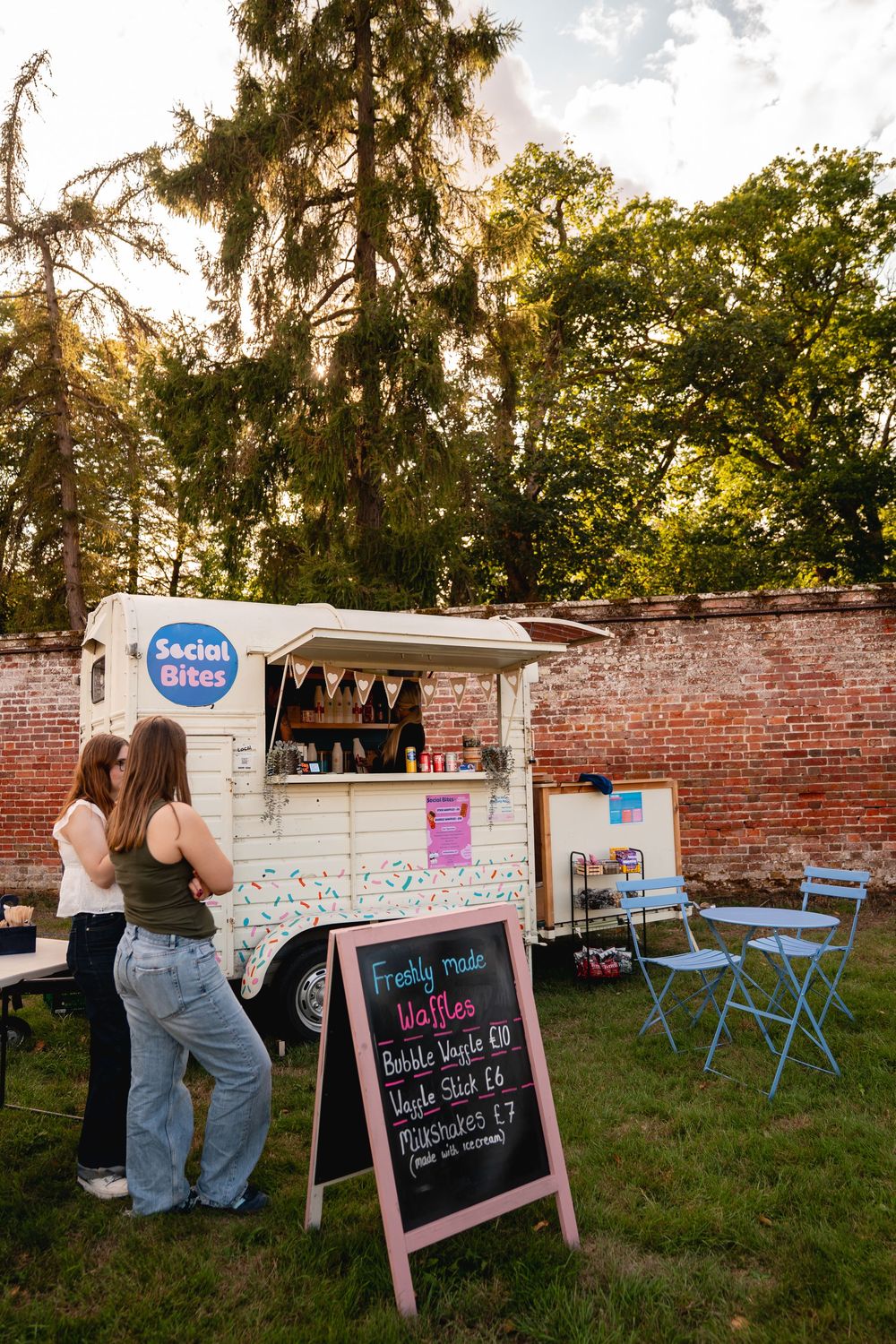 Two women ordering at Social Bites food truck serving freshly made waffles, parked outdoors near a brick wall.