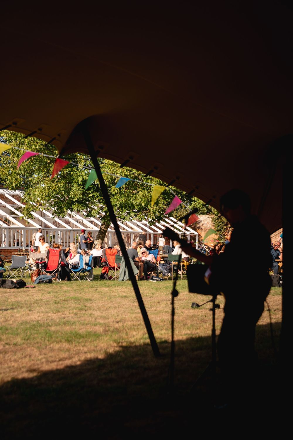 Silhouetted musician performs under a tent at an outdoor festival with colorful bunting and audience seated on grass.