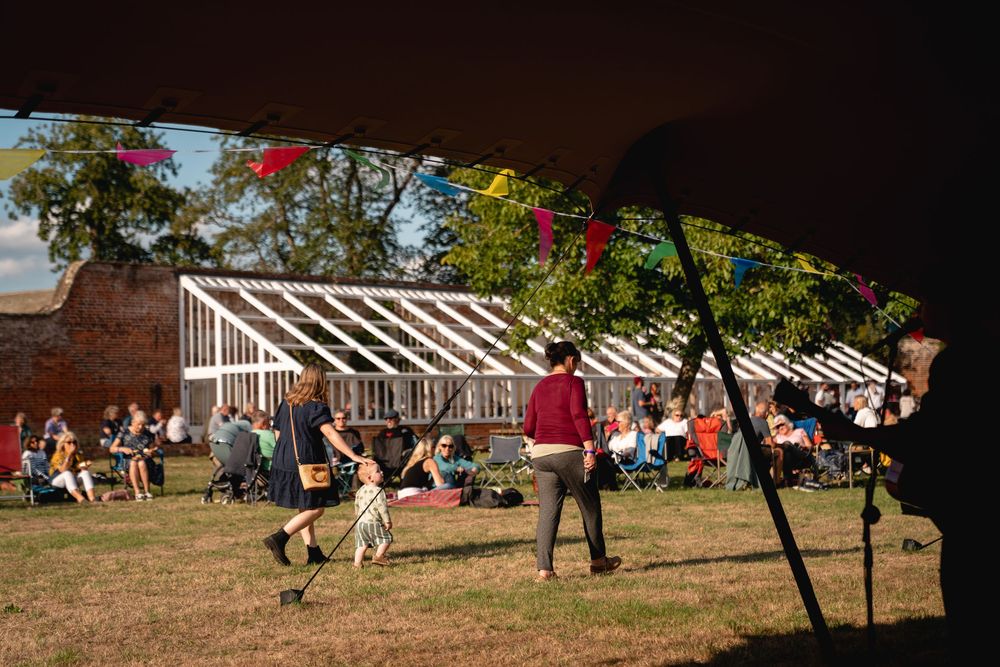 Outdoor festival gathering with people relaxing on grass near a white frame structure decorated with colorful bunting on a sunny day.