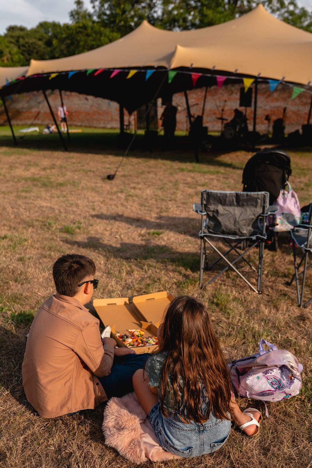 Couple sitting on grass sharing pizza at outdoor festival with colorful bunting and large tent in background.