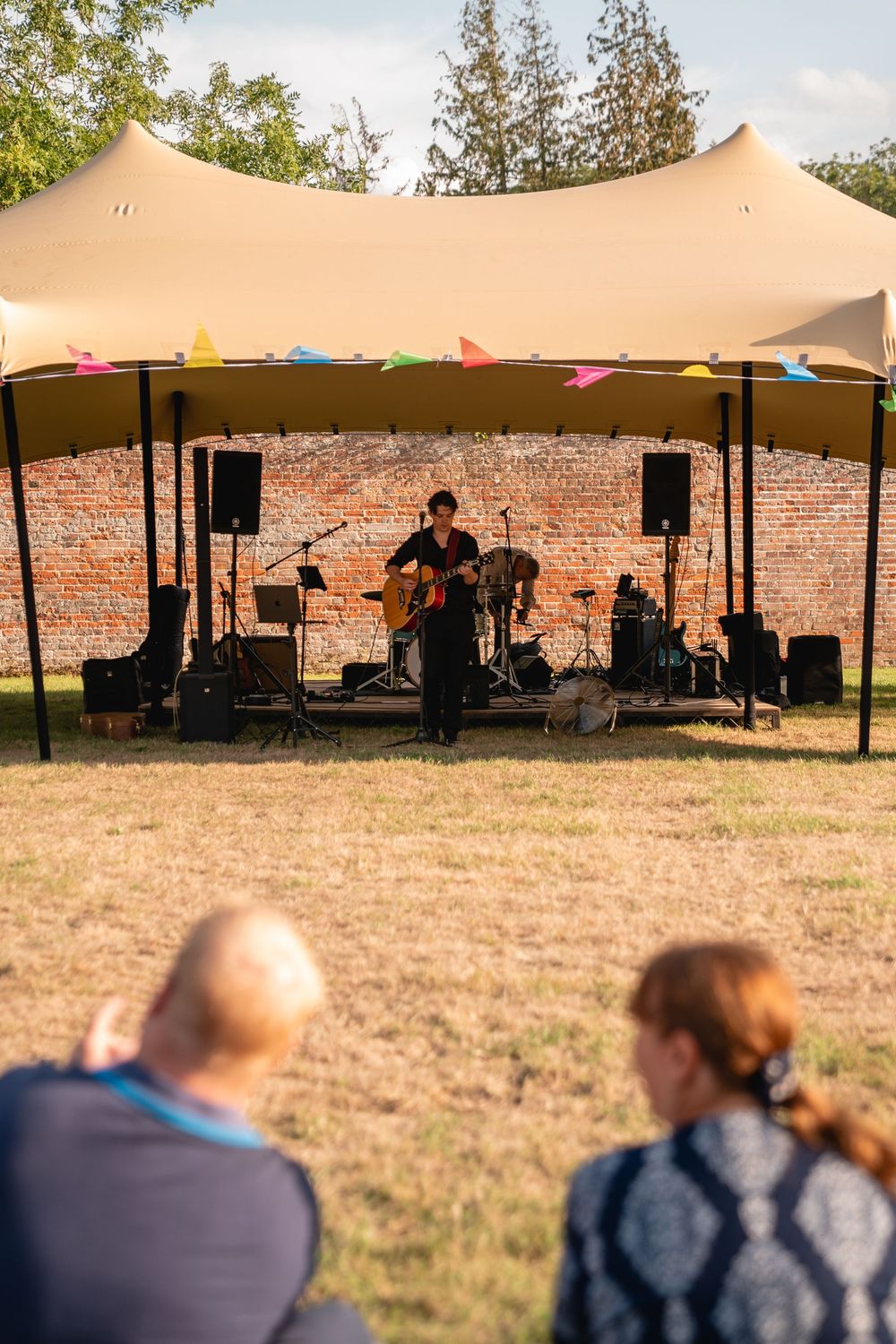 Outdoor music festival with guitarist performing on a tent-covered stage before a brick wall, watched by audience on grass.