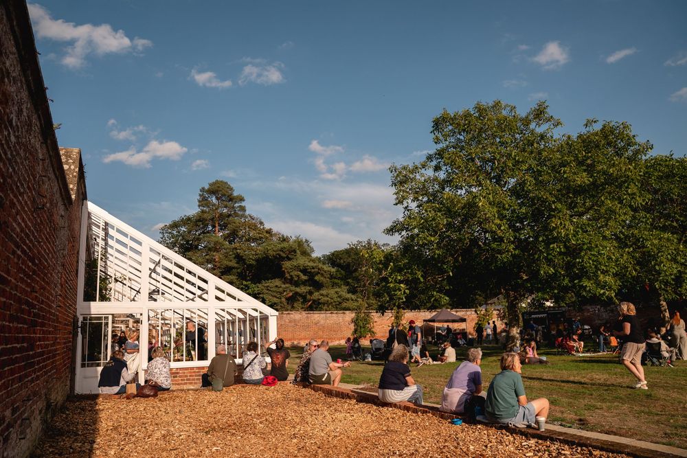 People relaxing outdoors on wood chips beside a white greenhouse structure in a sunny garden setting.