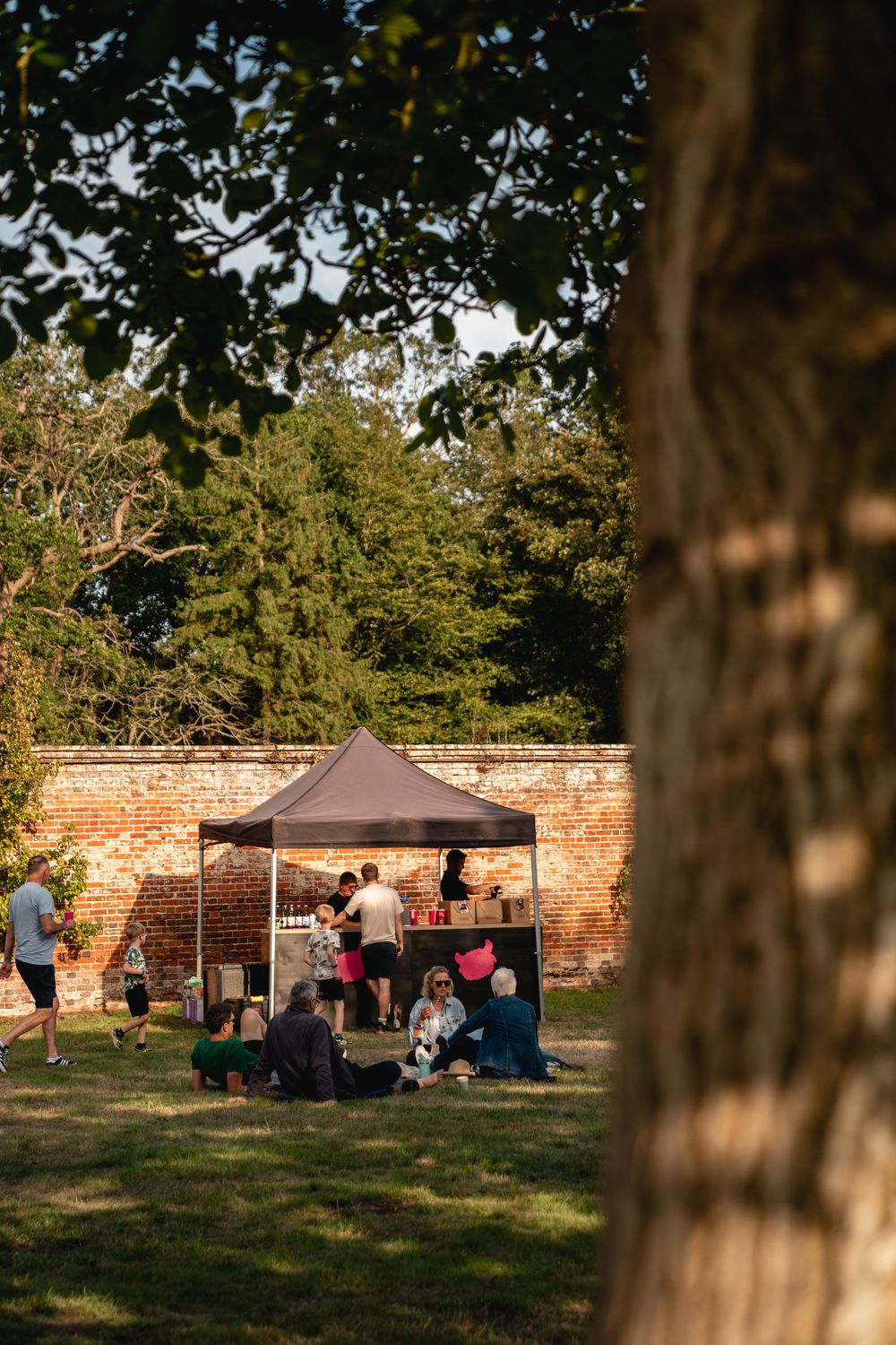 Outdoor market event with people gathered under a gazebo tent in a sunny walled garden surrounded by trees.