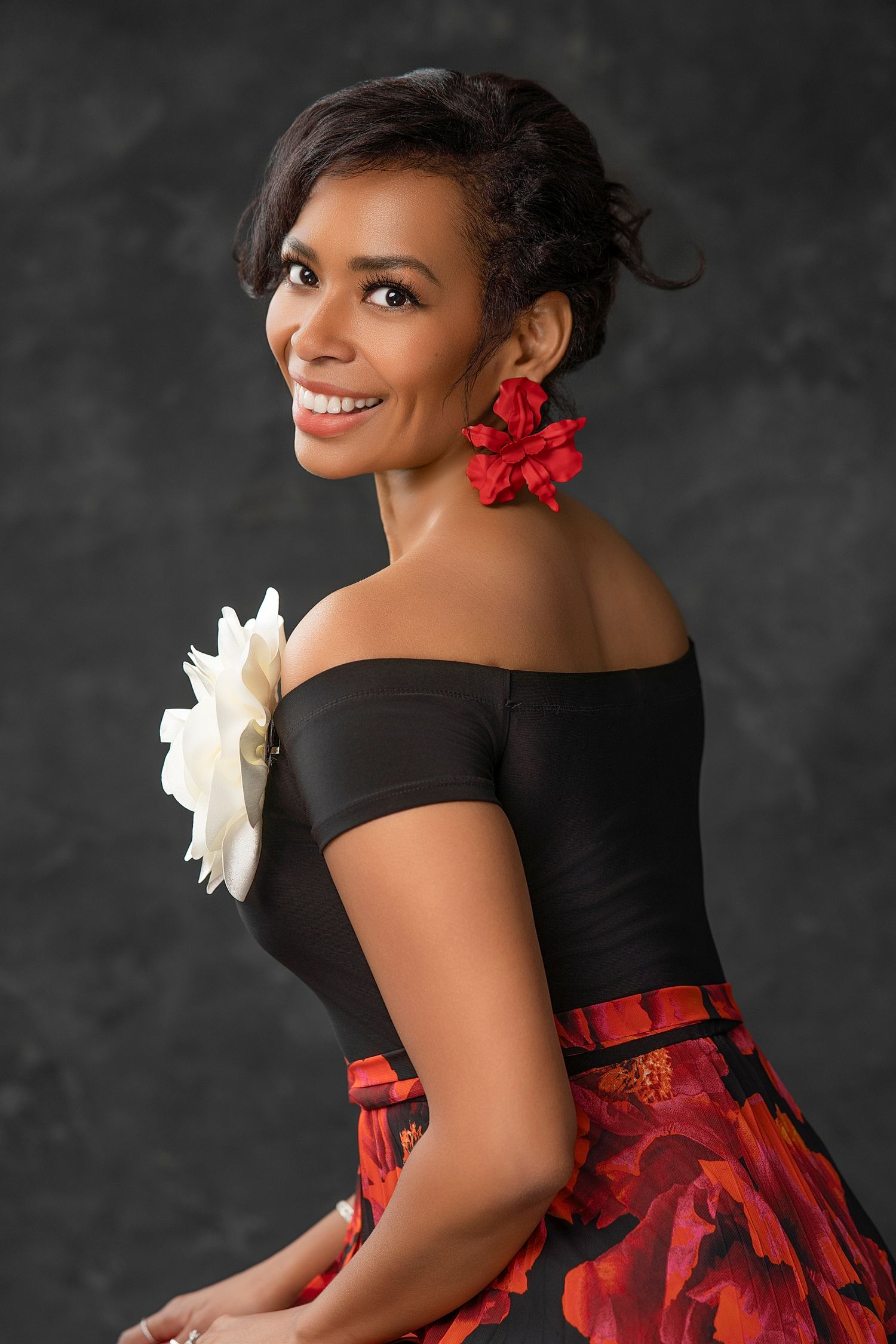 Smiling woman in off-shoulder black top with floral skirt, white flower brooch, and red flower earrings poses against gray backdrop.