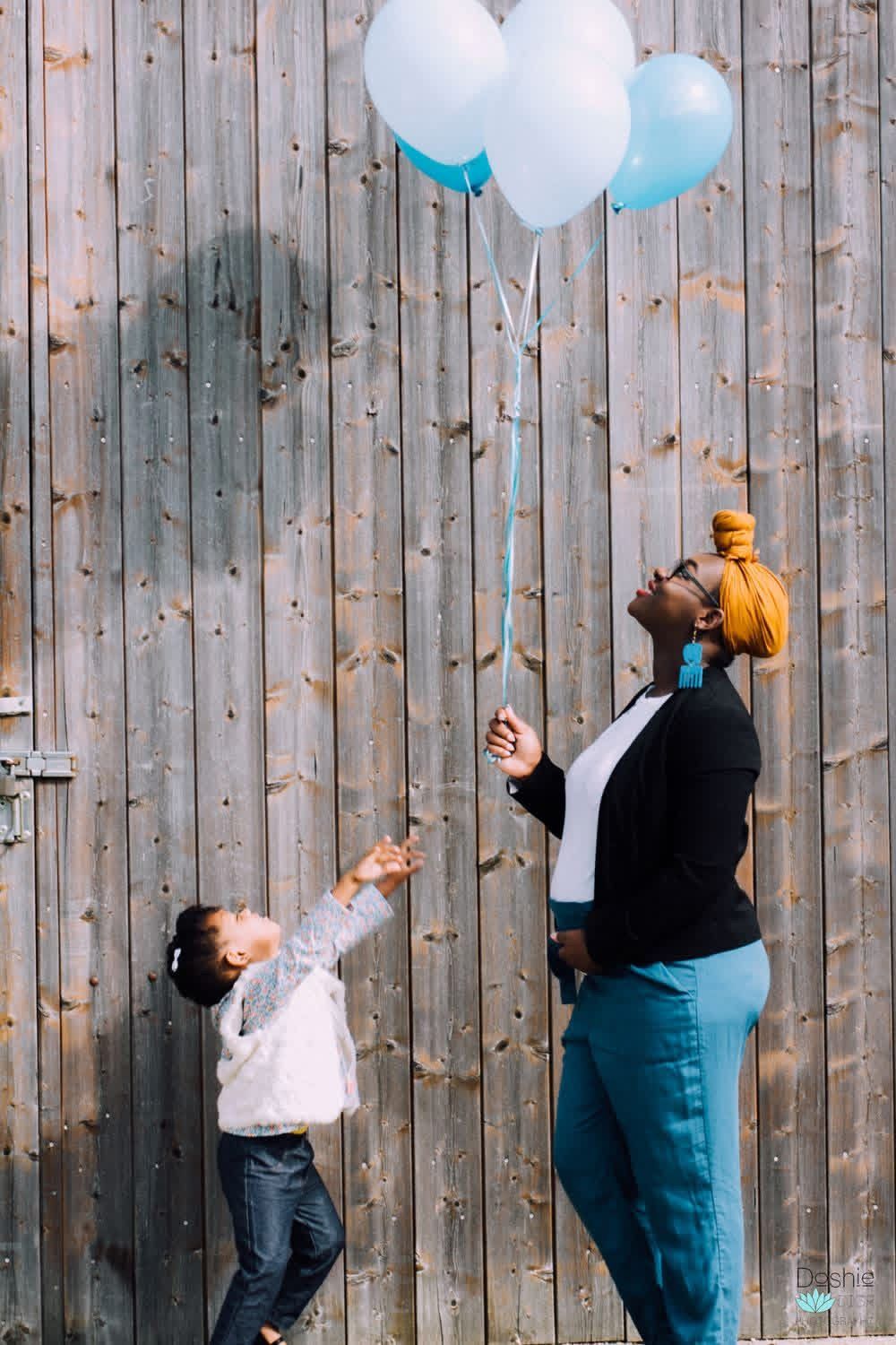 A woman and child look up at blue and white balloons against a rustic wooden wall outdoors.