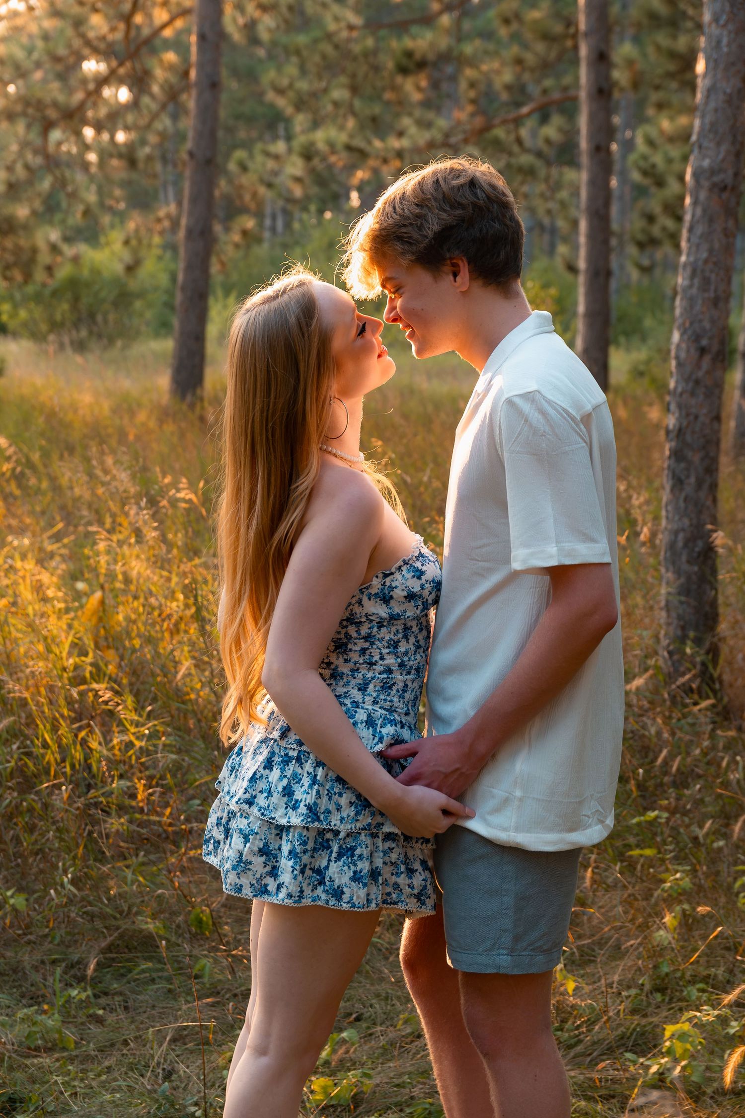 Young couple sharing a romantic moment in a sunlit forest, facing each other closely in summer outfits.