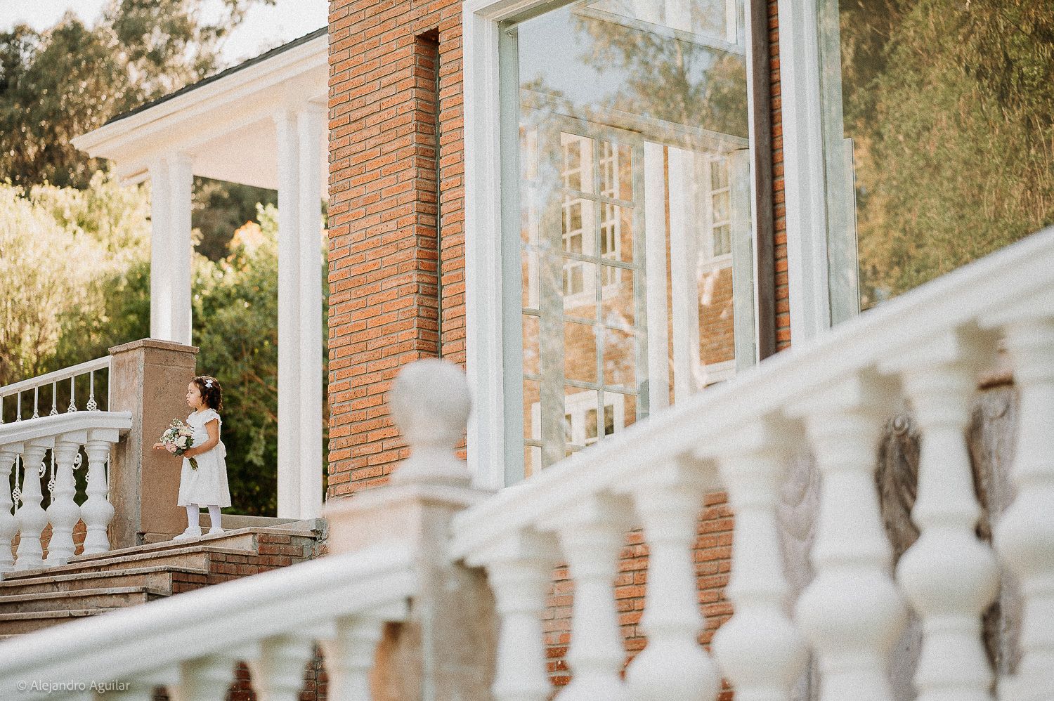 Bride holding bouquet stands on elegant white balustrade steps of a brick mansion with large glass windows.